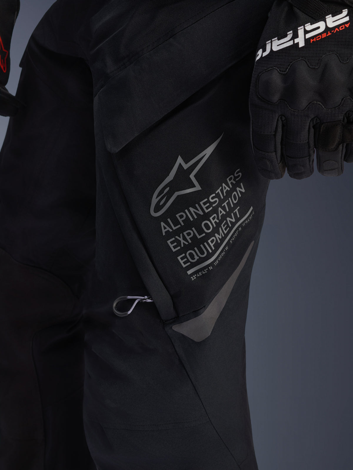 Primer plano de los pantalones Alpinestars ST-7 2L GORE-TEX en color negro, vista detallada del gran bolsillo cargo en el muslo con el distintivo gris de Alpinestars Exploration Equipment, el logo de la estrella y coordenadas geográficas, confección textil de alto rendimiento para adventure touring.