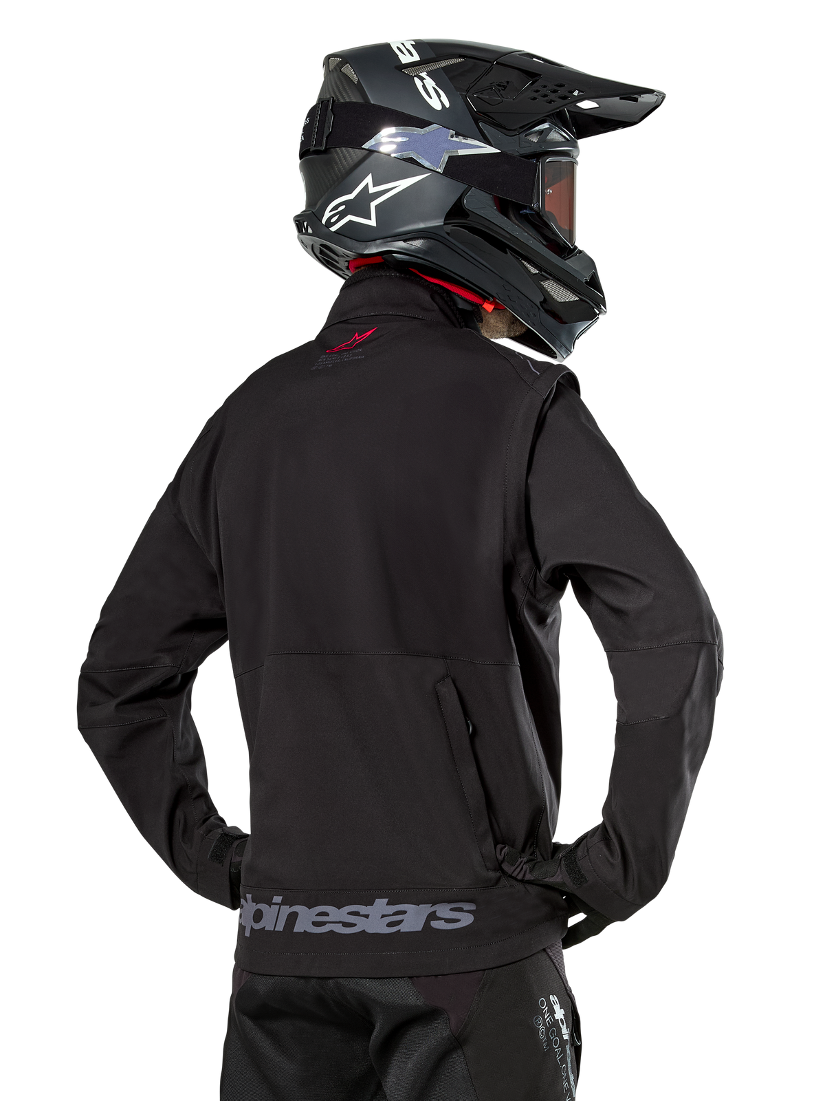 Chaqueta Softshell Alpinestars Lite-Dura, negra, vista trasera usada por un piloto, con marca Alpinestars gris en el dobladillo posterior y un pequeño logo rojo en el cuello, combinada con un casco de motocross negro, diseñada para enduro y conducción off-road