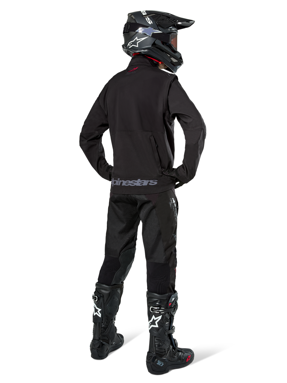 Piloto de enduro visto desde atrás con la chaqueta Alpinestars Lite-Dura Softshell en color negro, combinada con pantalones técnicos y botas negras, con un bolsillo de almacenamiento trasero con cremallera y el logotipo de Alpinestars en el dobladillo inferior, equipo completo de off-road para motocross.