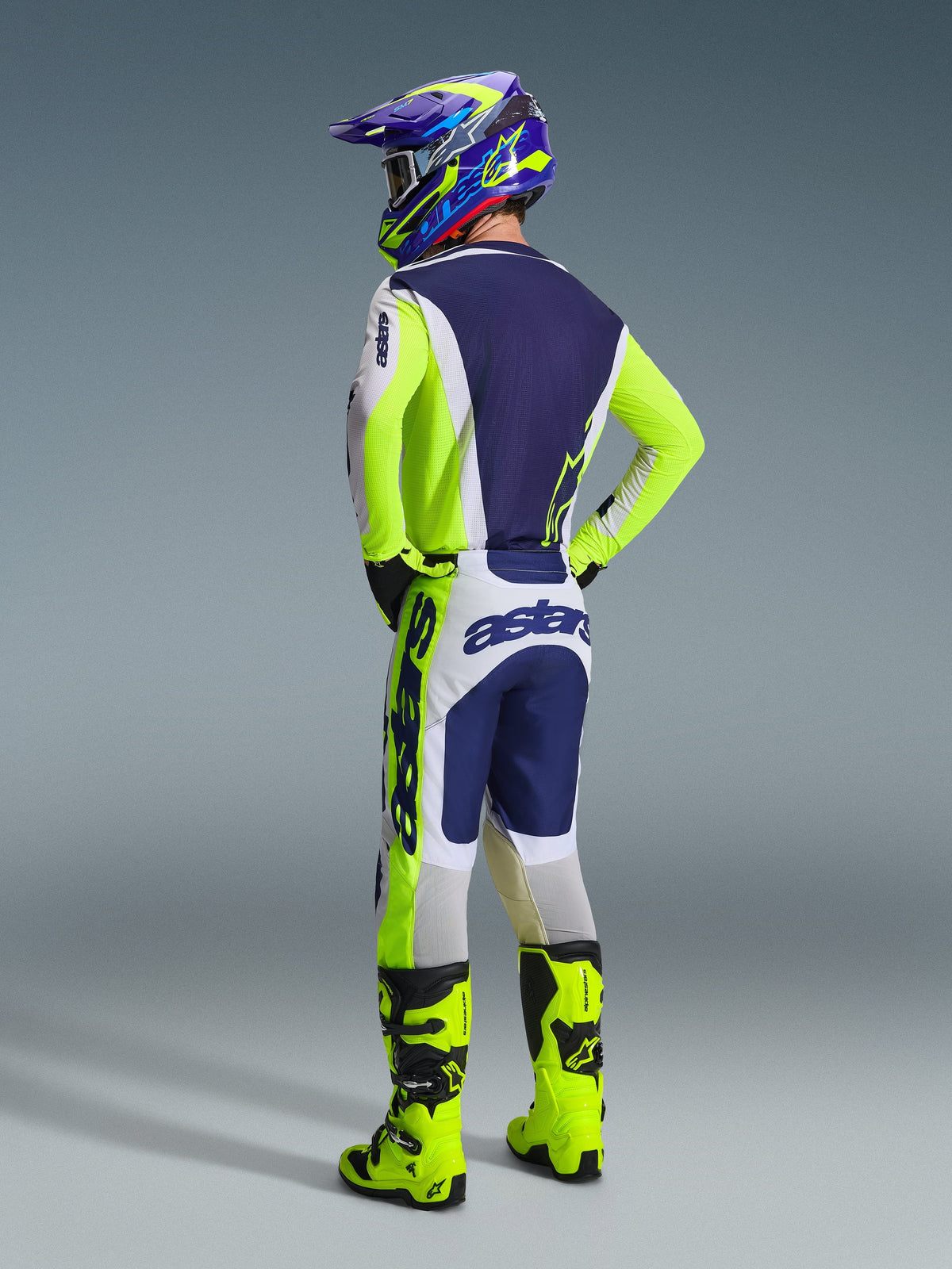 Motocross rider viewed from the back wearing complete racing gear, 2026 Racer Air Riway Pants in Light Gray, Yellow Fluo and Blue, matching jersey with navy blue back and neon yellow accents, coordinated helmet and neon yellow boots, professional motocross outfit.