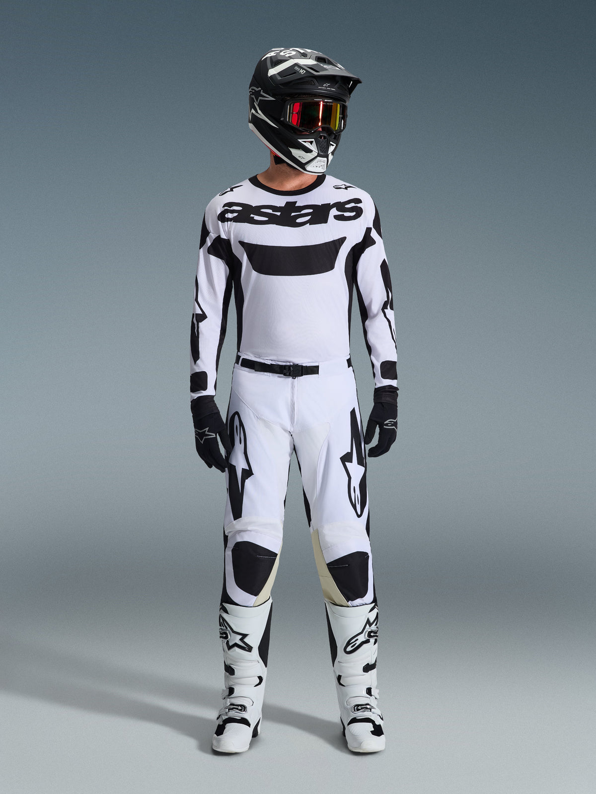 Piloto de motocross con pantalones Alpinestars Racer Riway 2026 en blanco y negro, combinados con un jersey blanco a juego, casco negro con gafas rojas, guantes negros y botas blancas, con logotipos Astars destacados en negro y un diseño de alto rendimiento para uso off-road.