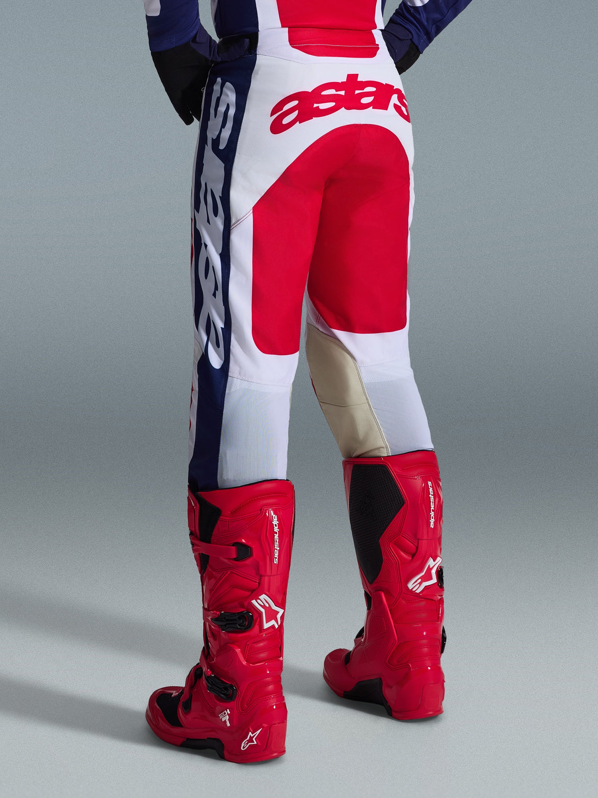 Alpinestars 2026 Racer Riway Pants, pantalones de motocross, blanco, azul y rojo, vista trasera que muestra un gran logotipo de Astars rojo en el asiento y paneles interiores de cuero en las rodillas de color canela, usados con botas de motocross Alpinestars rojas, equipamiento de rendimiento para carreras off-road