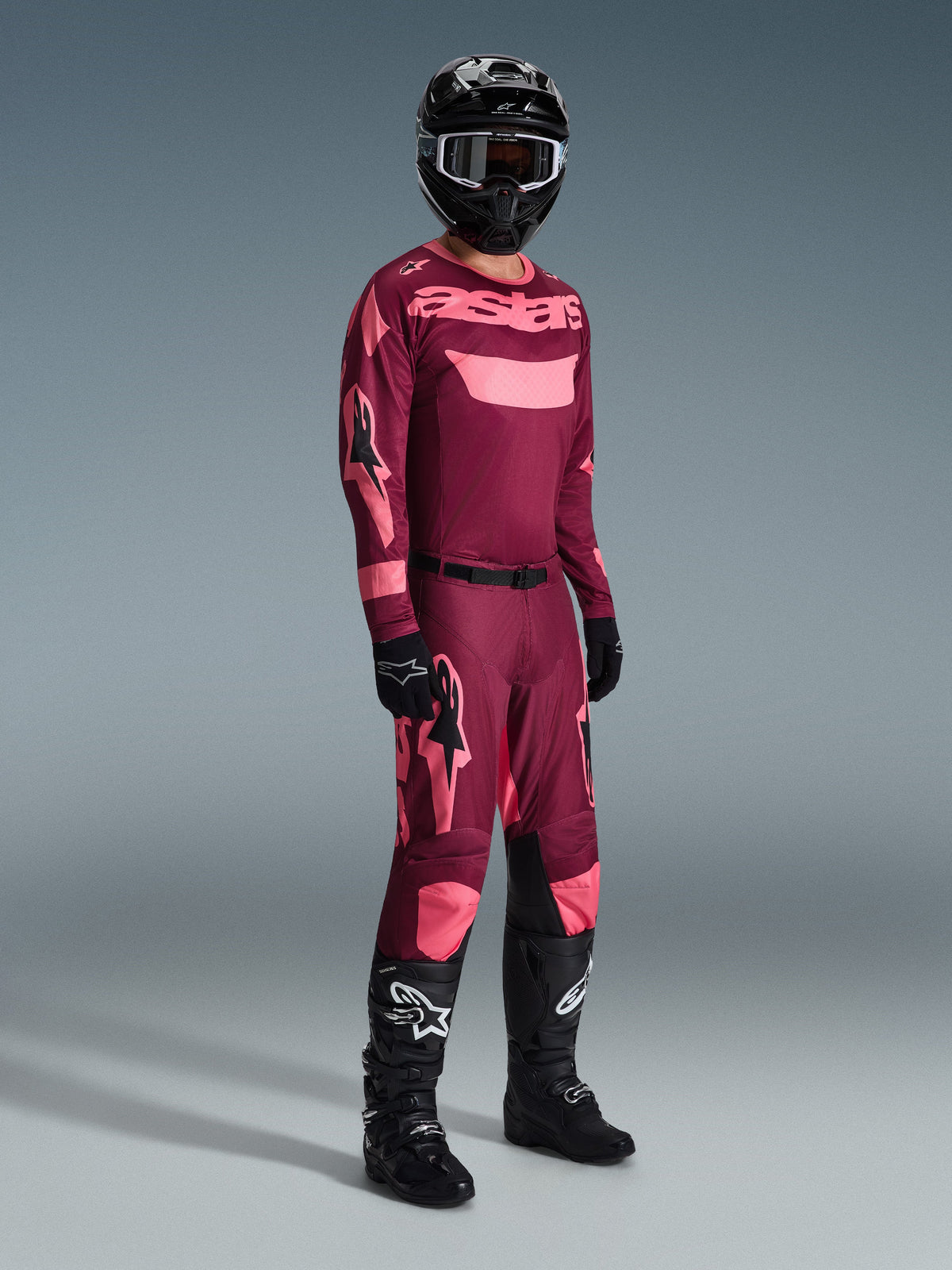 Alpinestars 2026 Racer Riway Gear Set, conjunto de jersey y pantalones de motocross en granate y rosa, con casco negro y botas negras de la serie Tech, vista de cuerpo completo sobre un fondo gris.