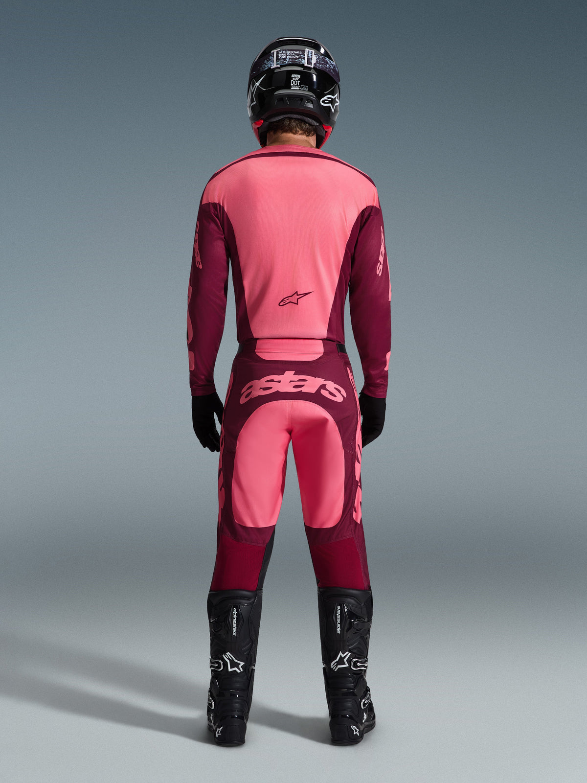 Vista trasera de un piloto de motocross con el conjunto de equipación Racer Riway 2026, jersey rosa y burdeos, pantalones burdeos a juego con el logotipo 'astars' en rosa claro en la parte trasera, casco negro, botas negras y guantes negros, atuendo coordinado para rendimiento off-road