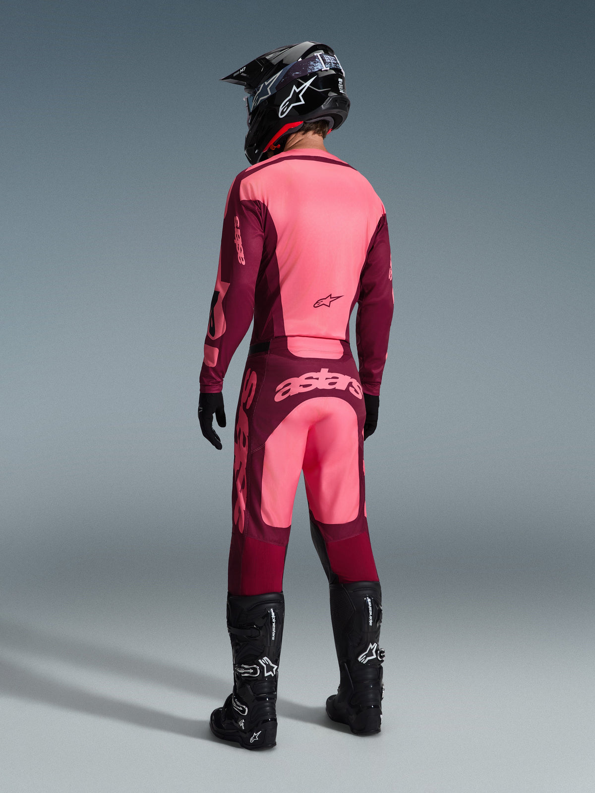Piloto de motocross con el conjunto de equipación Alpinestars 2026 Racer Riway visto desde atrás, combinación de colores granate y rosa claro, camiseta y pantalones con logotipos prominentes, completo con casco, guantes y botas negros, atuendo de carreras coordinado