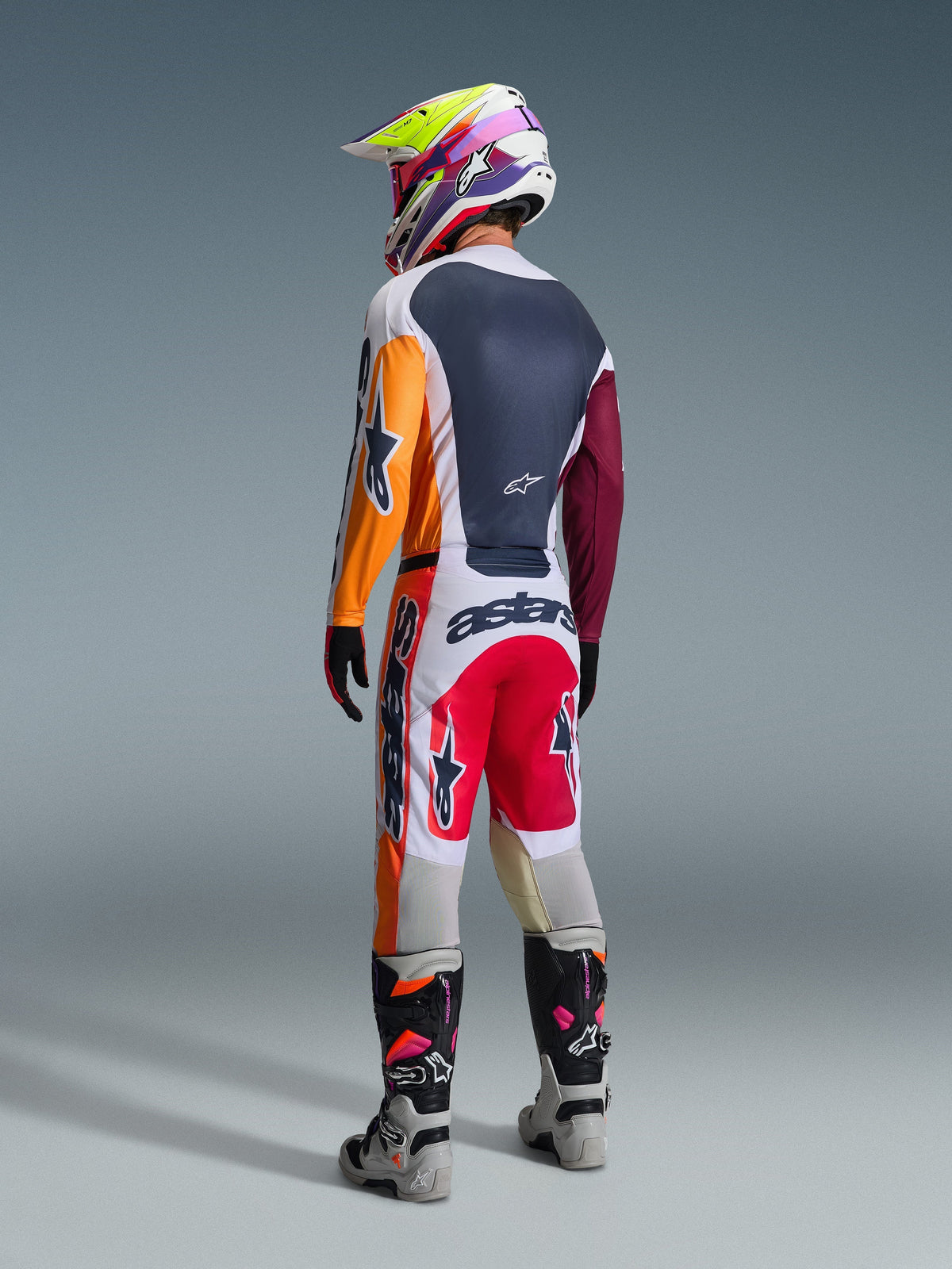 Motocross rider viewed from behind wearing complete racing gear, Alpinestars 2026 Racer Portl Pants in Red Light Gray and Dark Gray, matching jersey with orange and maroon sleeves, multi-colored graphic helmet, gray and black off-road boots with pink highlights, professional motocross outfit