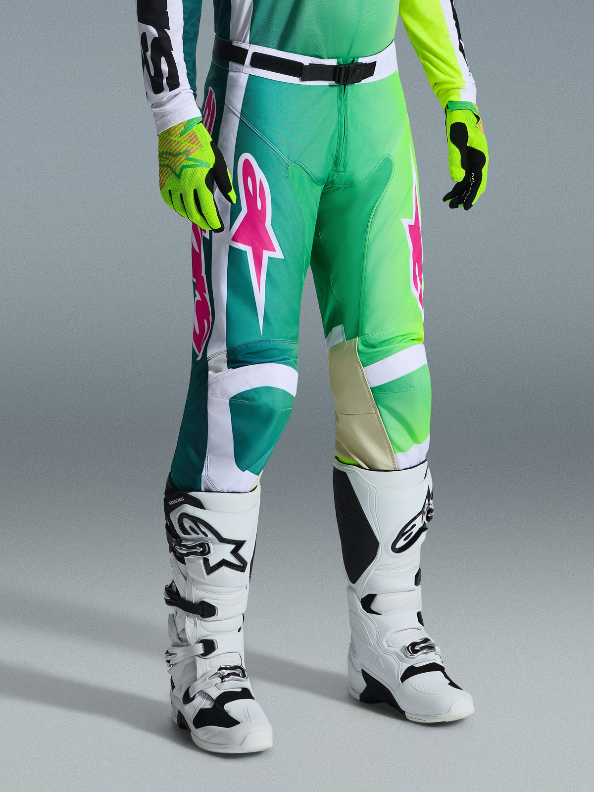 Alpinestars 2026 Racer Portl Pants, pantalones de motocross, verdes, blancos y negros con logotipos rosas, combinados con botas blancas y guantes amarillo neón, con llamativos patrones gráficos y hebilla de cintura ajustable negra, equipamiento de alto rendimiento para conducción todoterreno