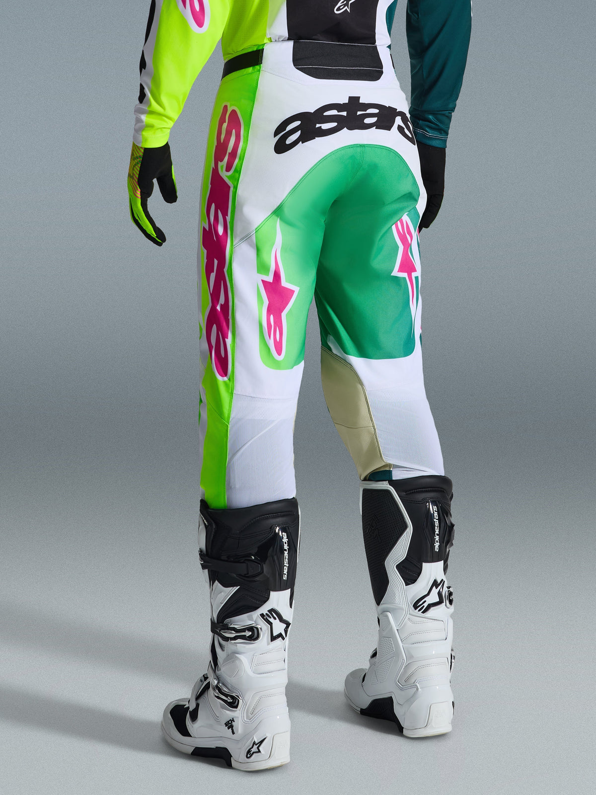 Alpinestars 2026 Racer Portl Pants, pantalones de motocross, verdes, blancos y negros con acentos en rosa, vista trasera con el llamativo logo 'astars' en el asiento y logos de estrella rosas en las piernas, combinados con botas de motocross blancas y negras, equipamiento de alto rendimiento para carreras off-road