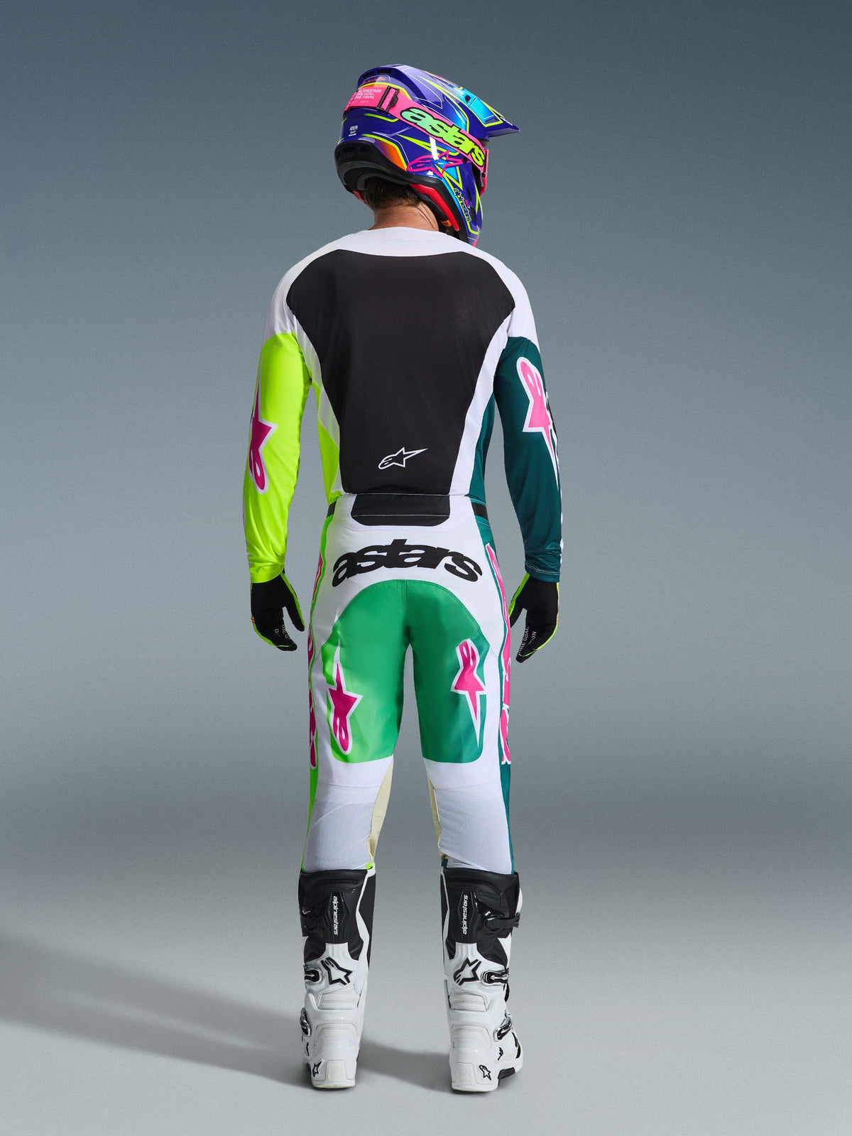 Piloto de motocross visto desde atrás con pantalones Alpinestars 2026 Racer Portl en verde, blanco y negro, jersey a juego con logotipos de estrellas rosas, casco multicolor 'astars' y botas blancas y negras, conjunto coordinado de equipamiento para carreras todoterreno.
