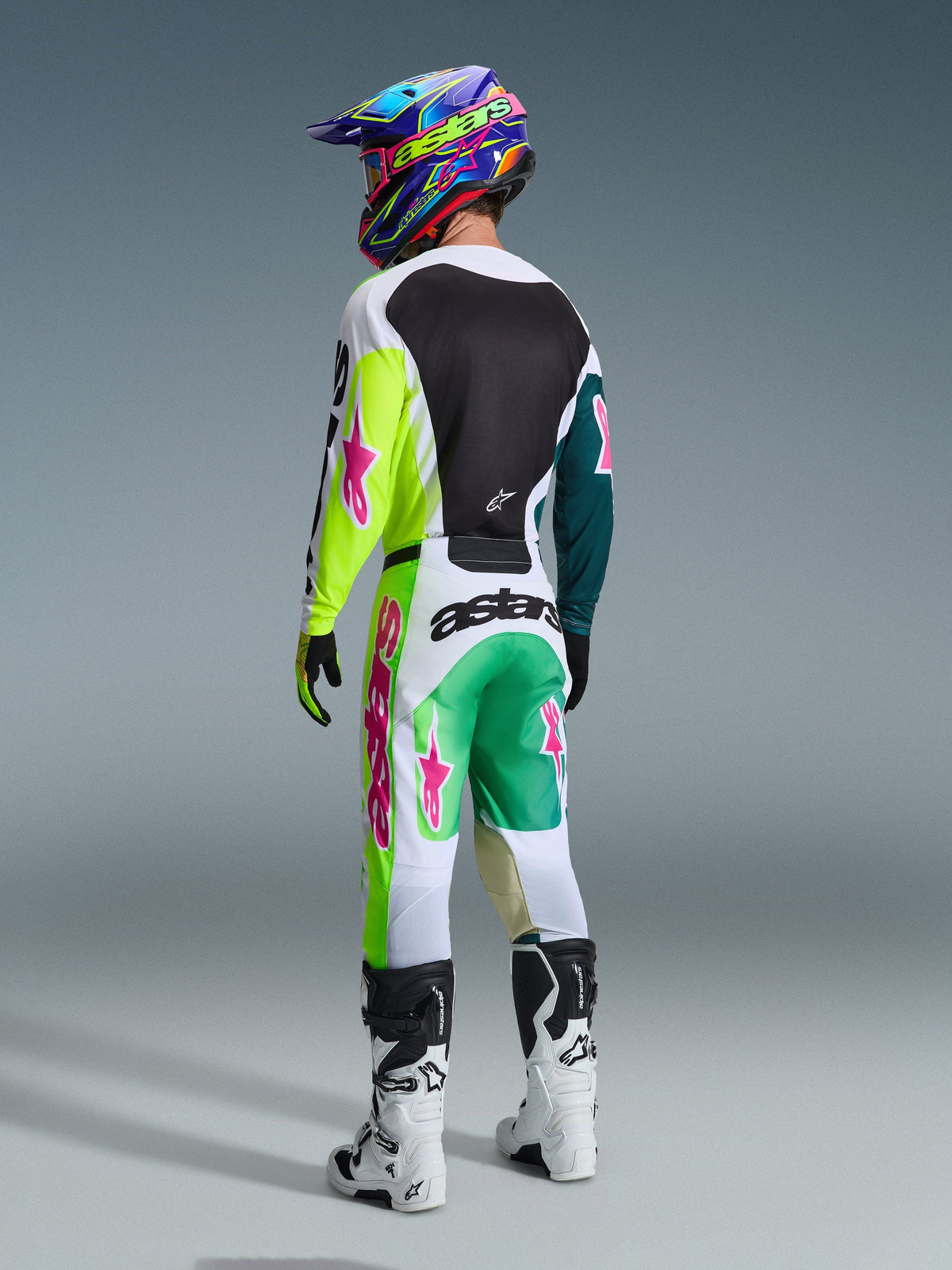 Piloto de motocross desde atrás con pantalones Alpinestars 2026 Racer Portl en verde, blanco y negro, jersey a juego con detalles en lima y rosa, casco colorido, botas blancas y negras, conjunto completo de equipamiento para carreras off-road.