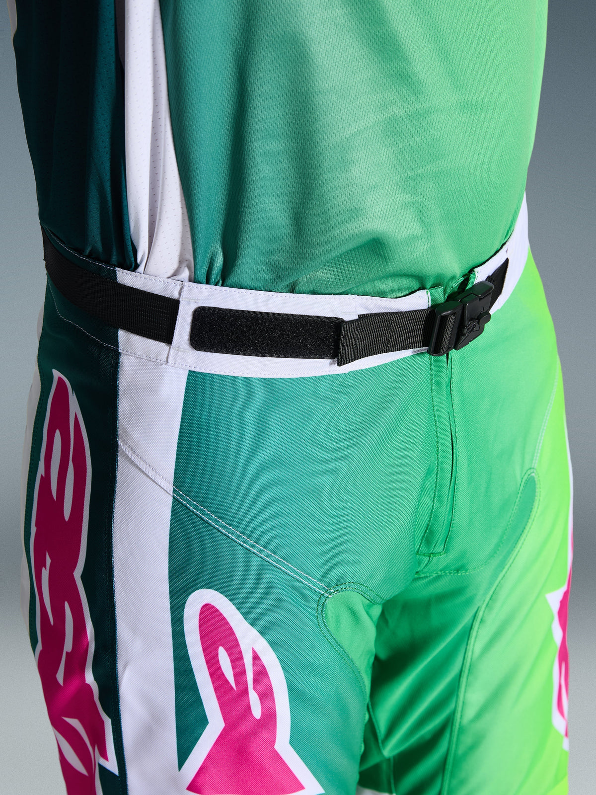 Alpinestars 2026 Racer Portl Pants, pantalones de motocross, verdes, blancos y negros con acentos del logo en rosa, primer plano de la cintura que muestra la hebilla magnética SX2 y el cierre ajustable, equipamiento duradero para carreras off-road
