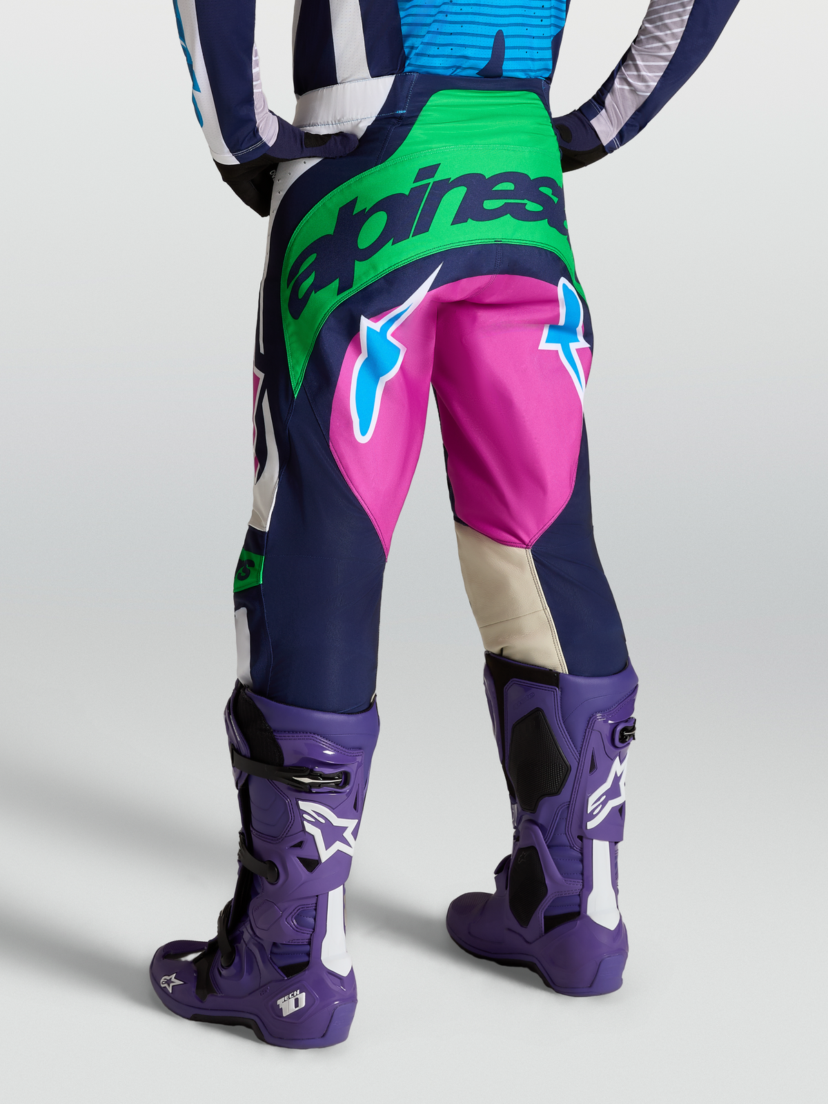 Alpinestars Techstar Maven, pantalones de motocross para hombre, azul y verde con detalles en rosa, vista trasera con botas Tech 10 moradas