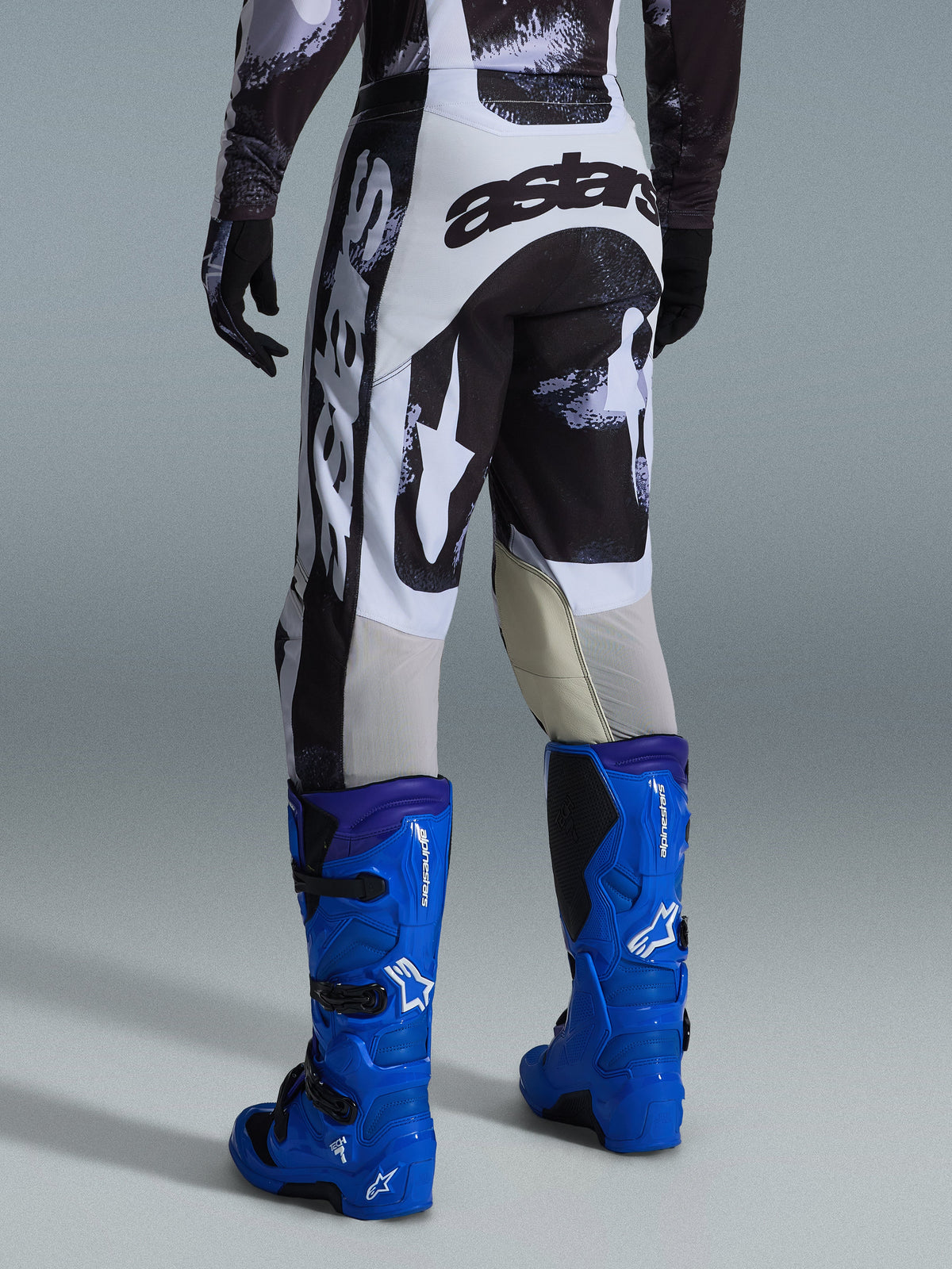 Pantalones Alpinestars Racer Lahnd 2026, pantalones de motocross, diseño Iron Camo gris y negro, vista posterior que muestra el logotipo astars grande en blanco y negro, combinados con botas de motocross azul brillante, equipamiento profesional de carreras off-road