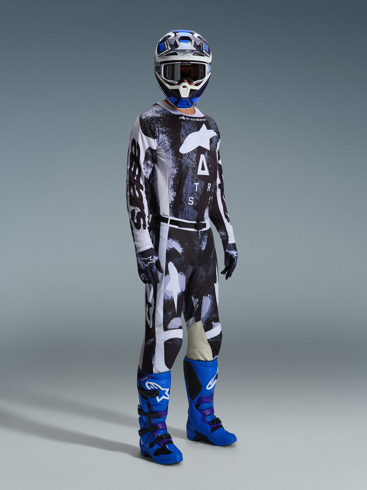 Piloto de motocross con equipamiento completo que incluye los pantalones Alpinestars 2026 Racer Lahnd, con patrón Iron Camo y gris, jersey a juego, botas de motocross azul brillante y púrpura, casco azul y blanco, conjunto profesional para carreras off-road