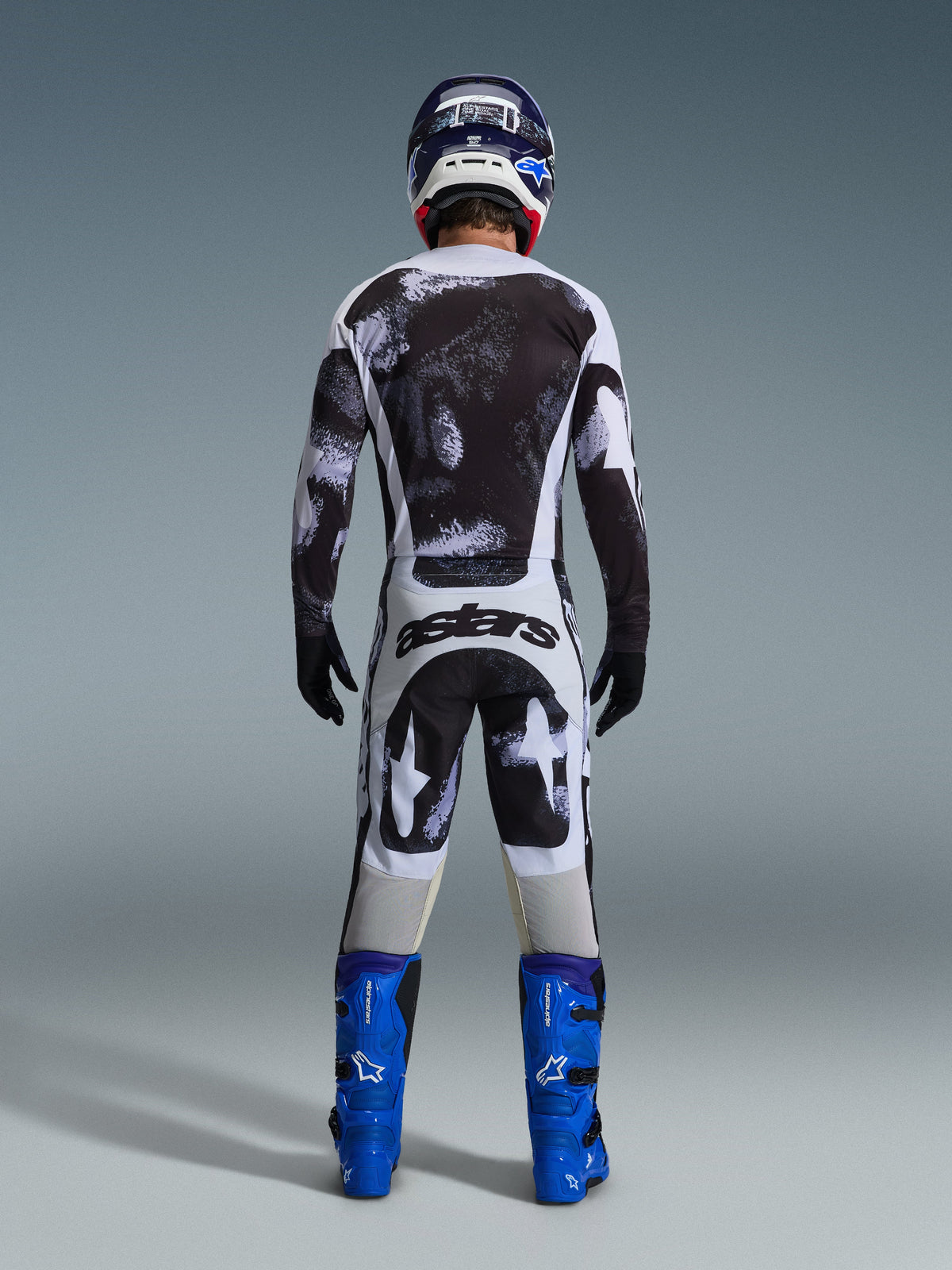 Alpinestars 2026 Racer Lahnd Pants, pantalones de motocross, patrón de camuflaje Iron Camo en gris y negro, vista trasera de un piloto con equipamiento completo de carreras, jersey a juego y botas azules, diseñados para rendimiento off-road