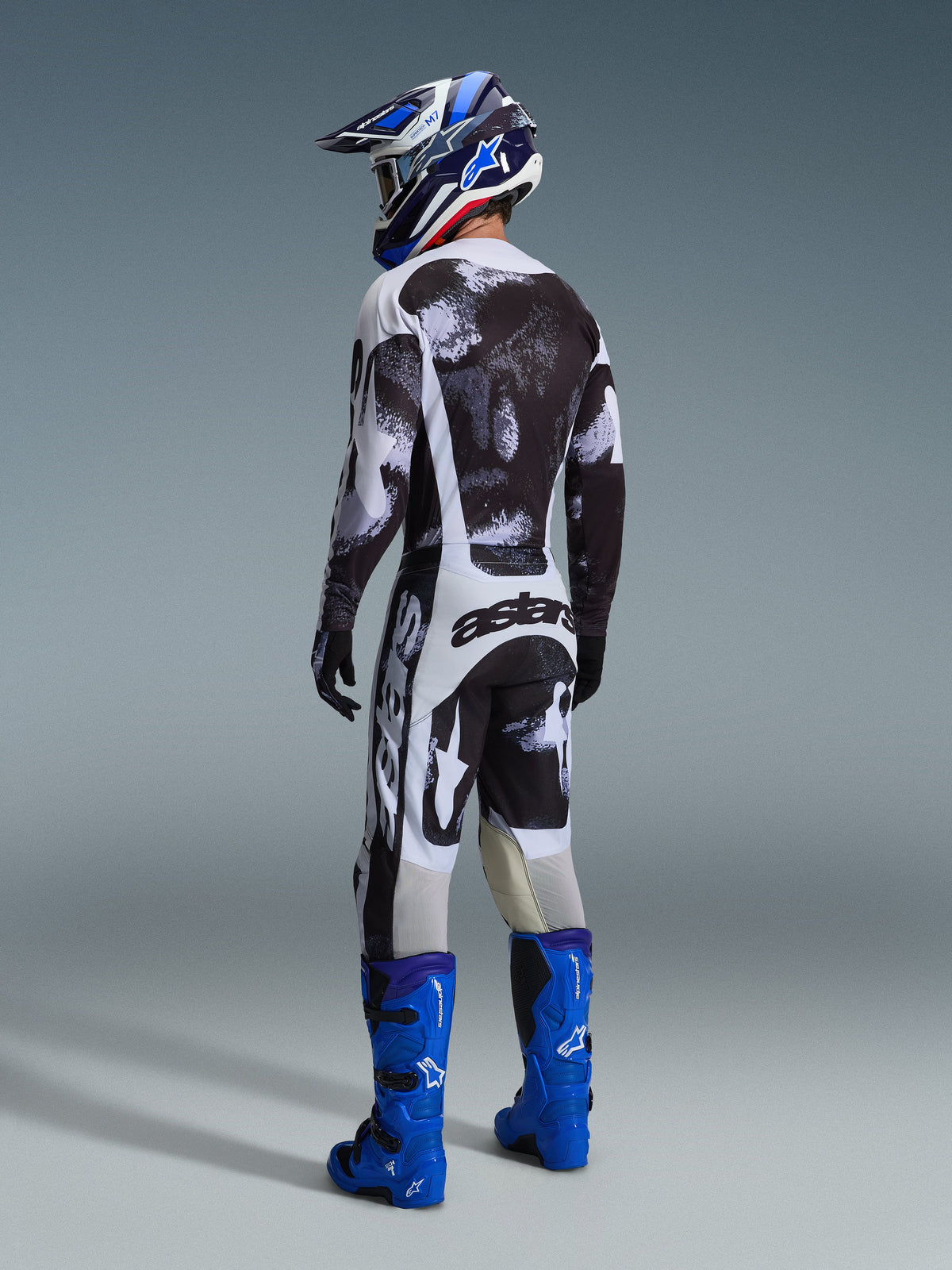 Piloto de motocross visto desde atrás con pantalones Alpinestars 2026 Racer Lahnd en Iron Camo, jersey con patrón en blanco y negro a juego, botas técnicas azul brillante y casco azul y blanco, equipo de carreras off-road coordinado con marca 'astars' llamativa en la parte trasera.