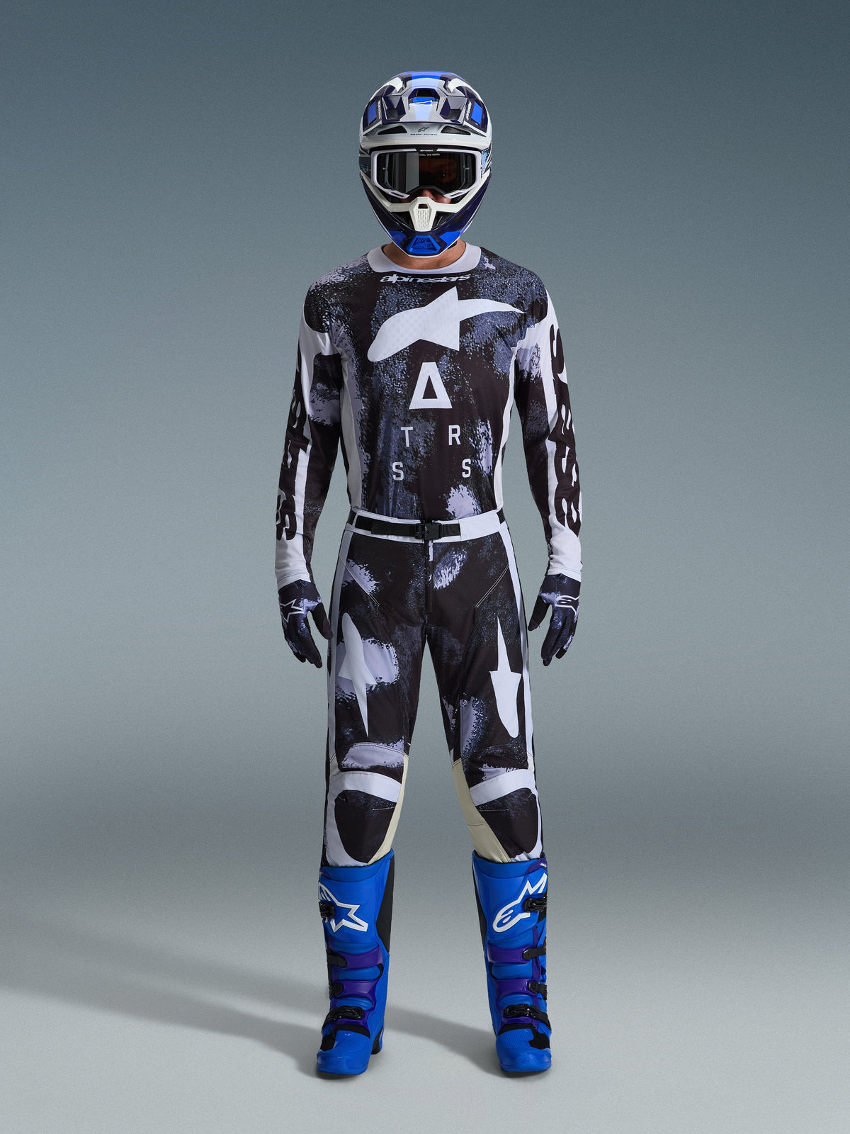 Piloto de motocross vistiendo los pantalones Alpinestars 2026 Racer Lahnd en Iron Camo, diseño de camuflaje gris y negro, jersey a juego, casco azul y blanco con gafas y botas azul brillante, conjunto de equipamiento de motocross coordinado.