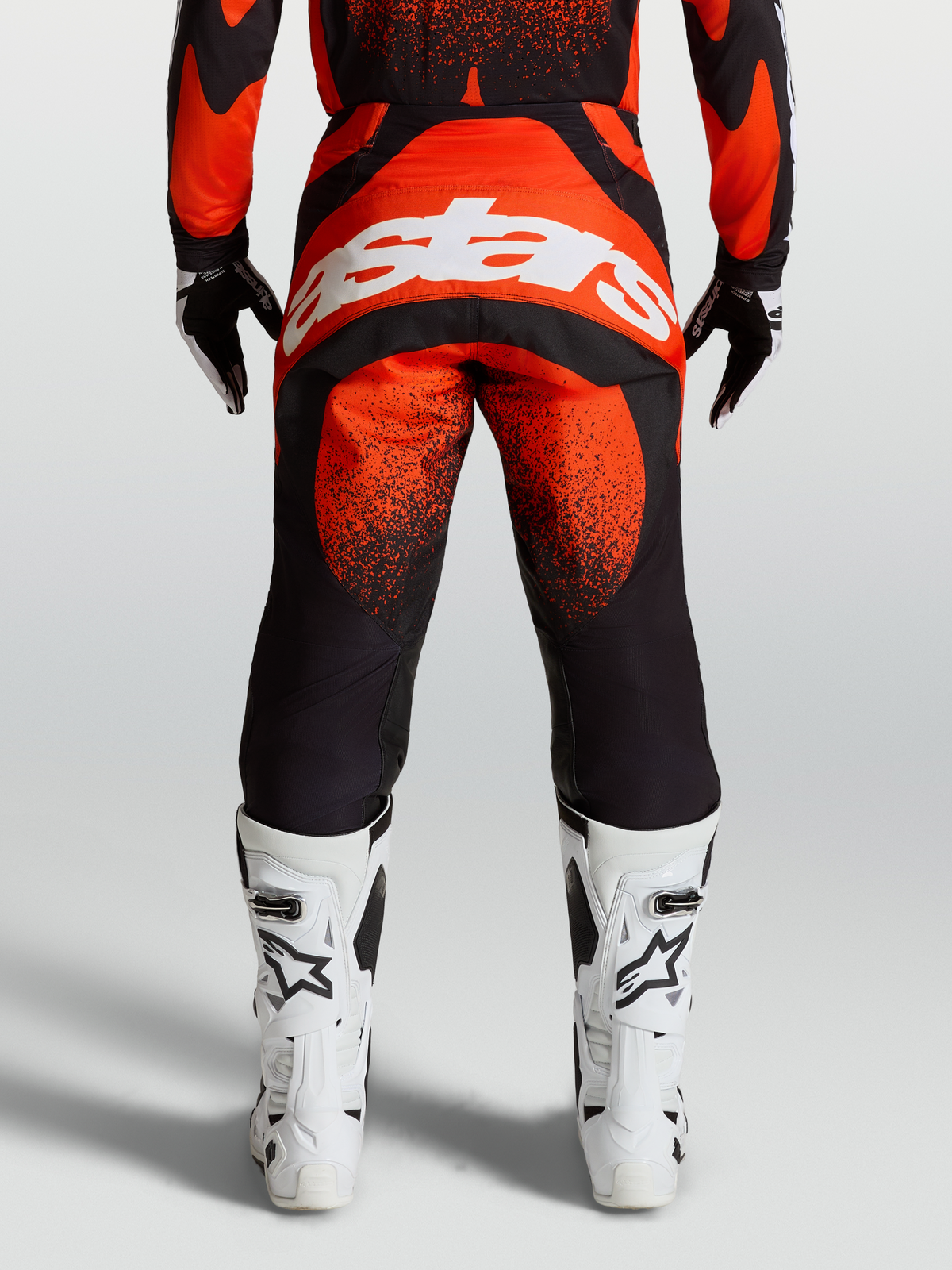 Alpinestars Techstar Swan, pantalones de motocross para hombre, rojos y negros con logo blanco en el asiento, vista trasera en modelo con botas blancas de carreras
