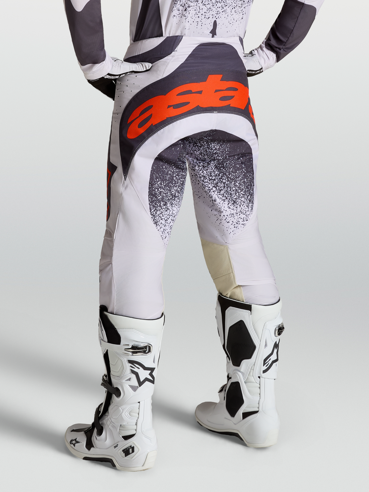 Alpinestars Pantalones Techstar Swan, pantalones de motocross para hombre, lila claro y gris oscuro con detalles en rojo flúor, usados con botas Tech 10 blancas.