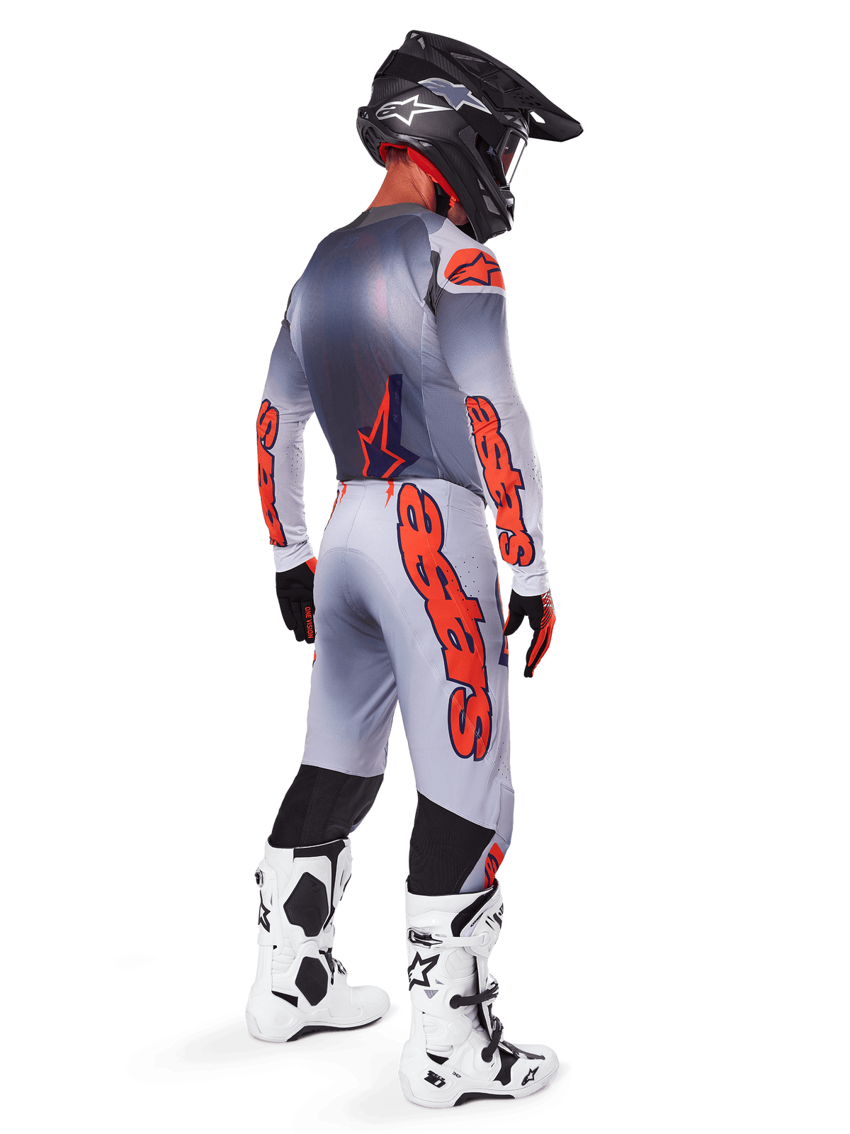 Piloto de motocross visto desde atrás vistiendo pantalones Alpinestars 2025 Supertech Lipan en gris y naranja flúor, jersey de manga larga a juego, casco negro y botas técnicas blancas y negras, equipamiento profesional completo de carreras off-road.
