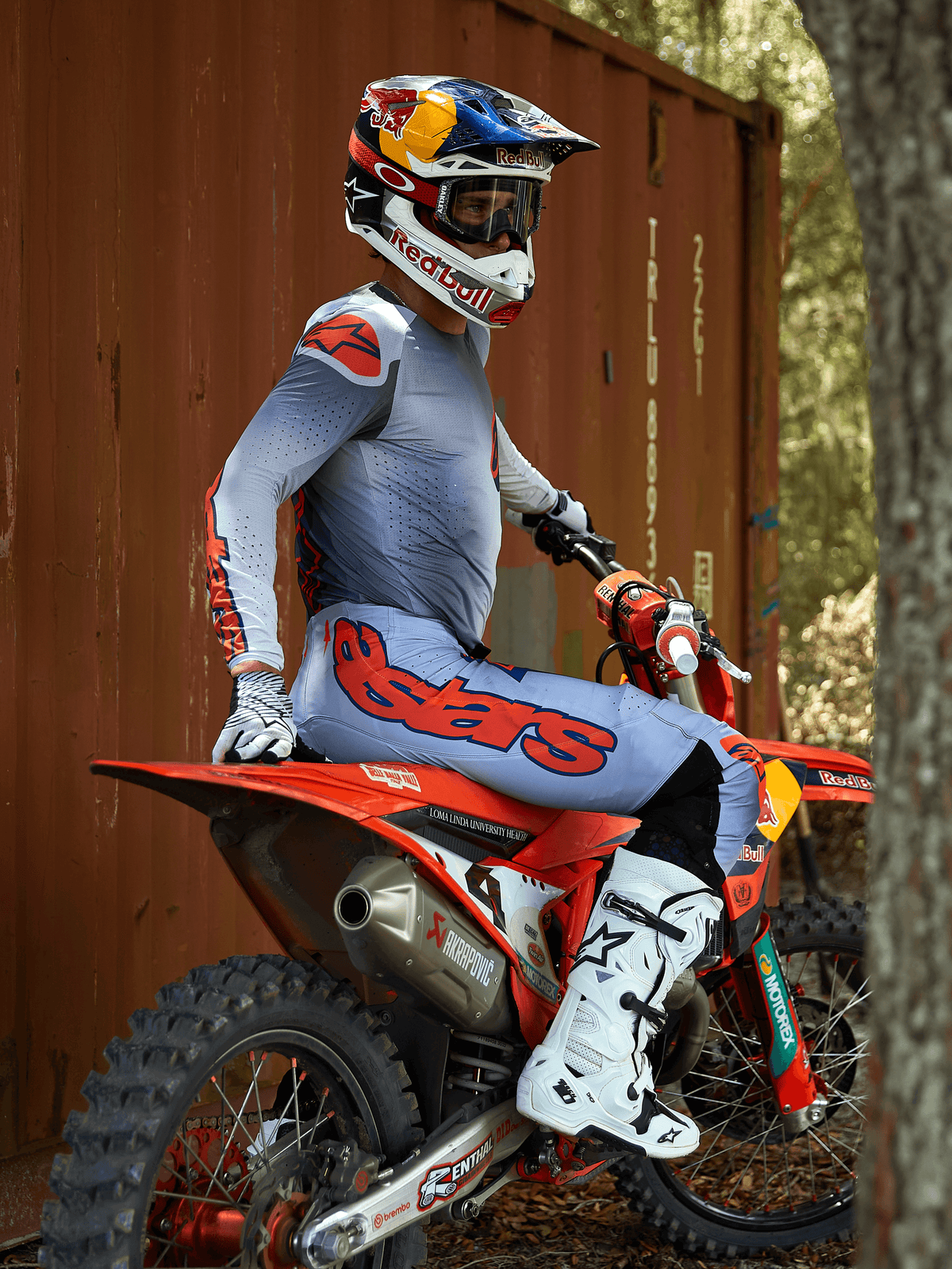Piloto de motocross sentado en una moto de cross roja, vistiendo el conjunto Alpinestars 2025 Supertech Lipan en gris y naranja flúor, pantalones azul claro con un llamativo logotipo de Astars en naranja, jersey a juego, botas de motocross blancas y un casco de Red Bull, equipamiento profesional de competición coordinado