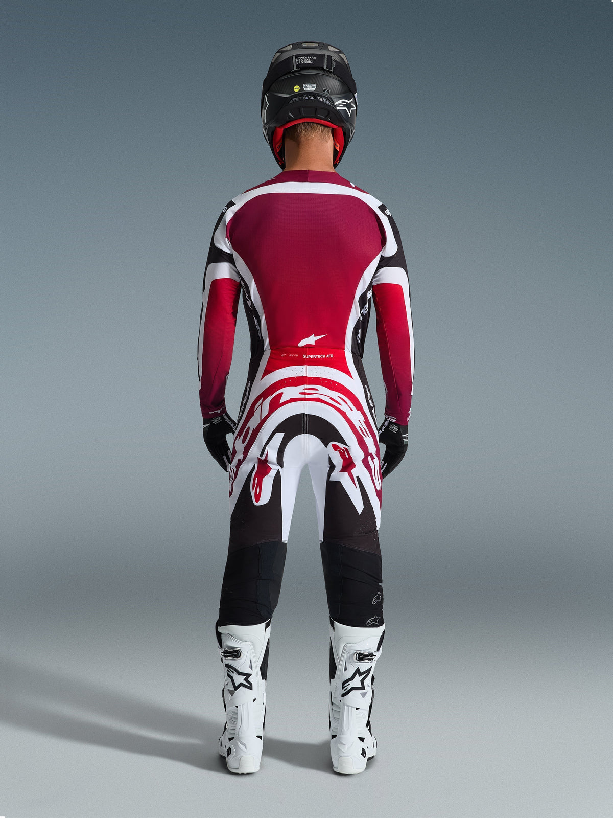 Piloto de motocross visto desde atrás luciendo los pantalones Alpinestars 2026 Supertech AFD Vista, negro, rojo y blanco, combinados con un jersey a juego, botas de competición blancas y casco negro; conjunto de equipamiento profesional completo para carreras off-road