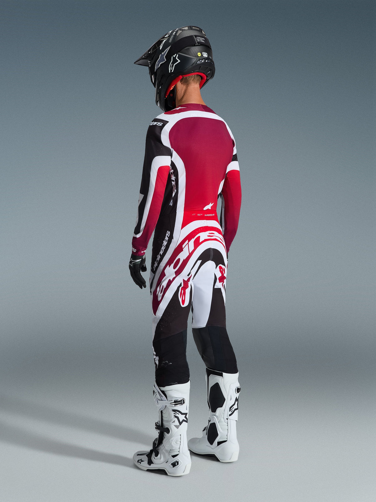 Piloto de motocross desde atrás vistiendo pantalones Alpinestars Supertech AFD Vista 2026, negro, rojo y blanco, con jersey a juego, casco negro y botas blancas y negras, vista trasera de cuerpo completo de equipamiento de competición profesional