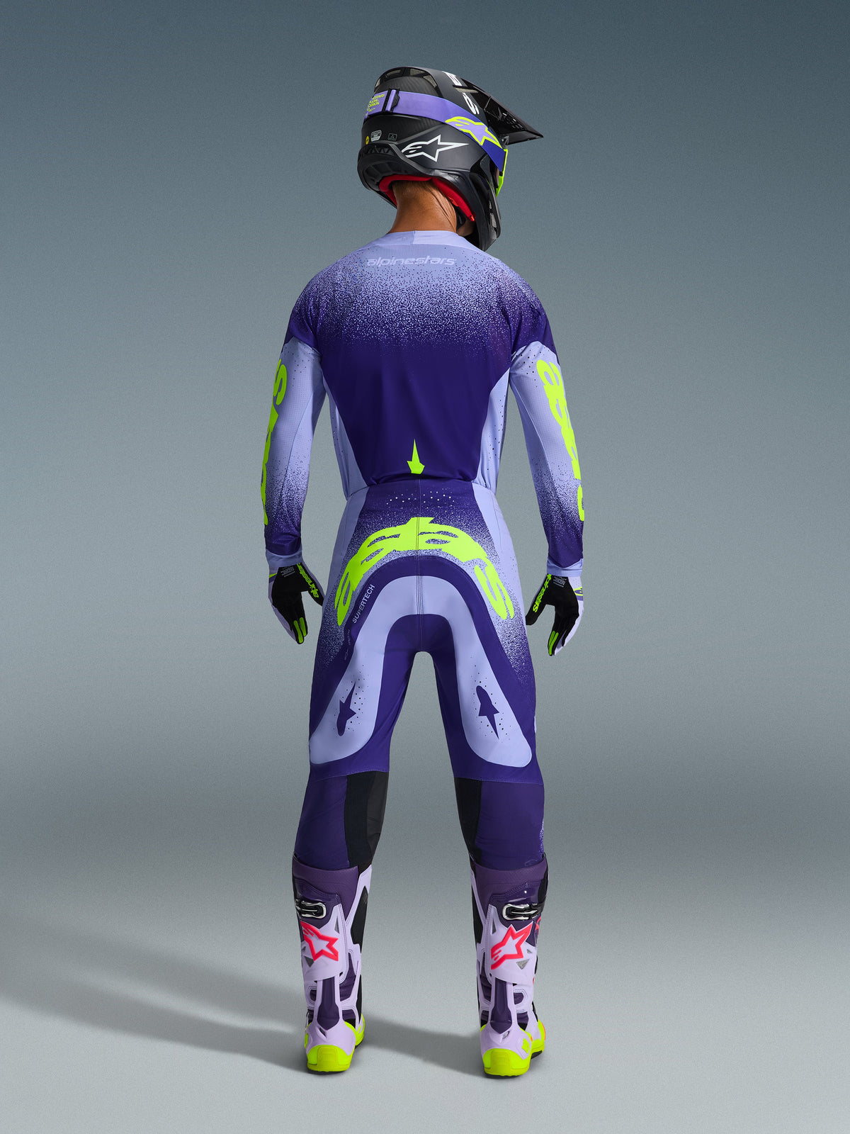 Motocross rider from behind wearing Alpinestars 2026 Supertech Scenz Pants in Purple Yellow Fluo, matching jersey with gradient purple and neon yellow graphics, black helmet, and purple boots, coordinated professional racing gear for off-road performance