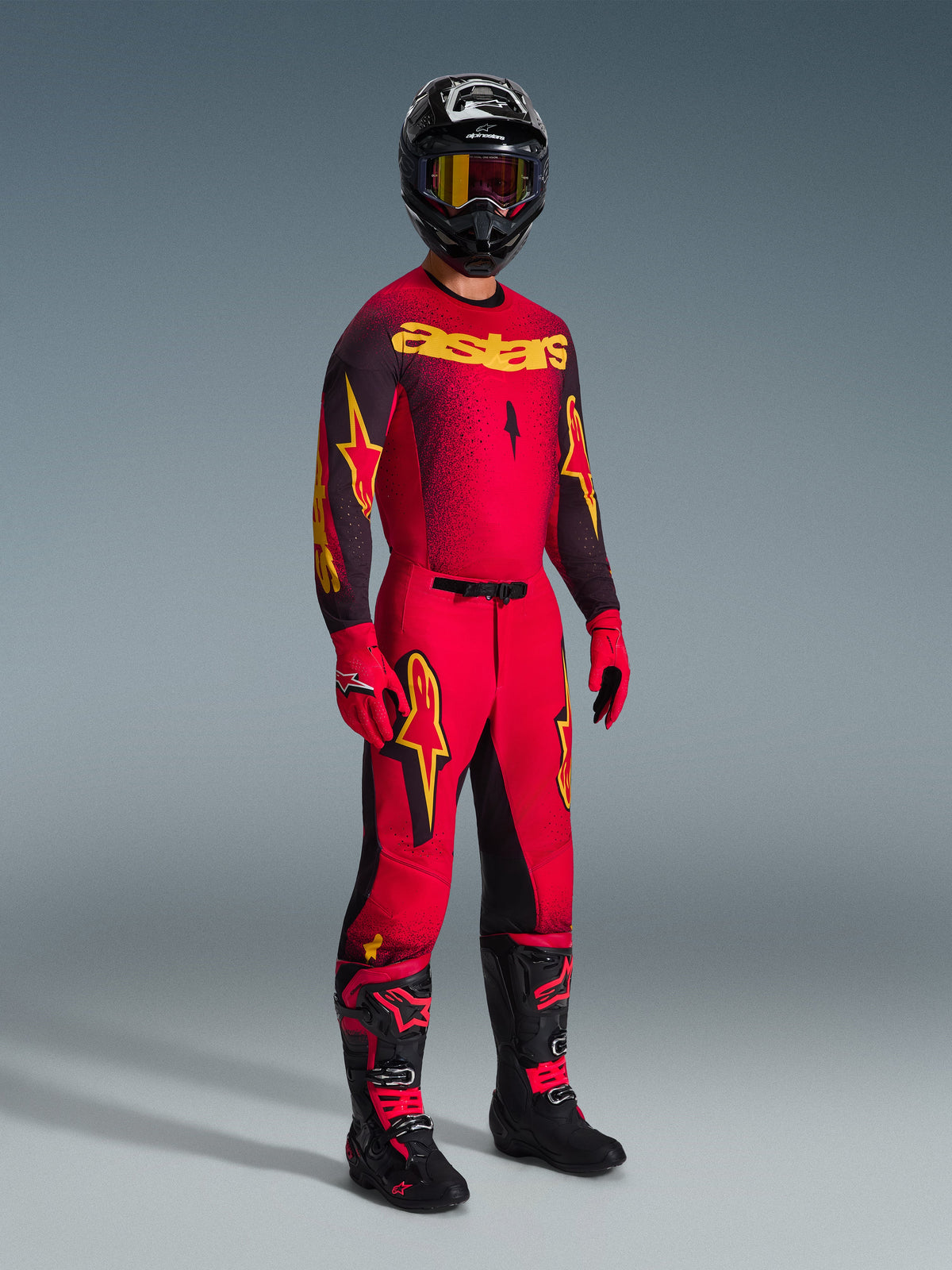 Alpinestars 2026 Supertech Scenz Pants, pantalones de motocross, rojos, negros y amarillos, usados por un piloto en un conjunto completo de equipamiento de carreras con jersey y botas a juego, con llamativos logotipos de estrellas en amarillo y negro y un ajuste atlético.