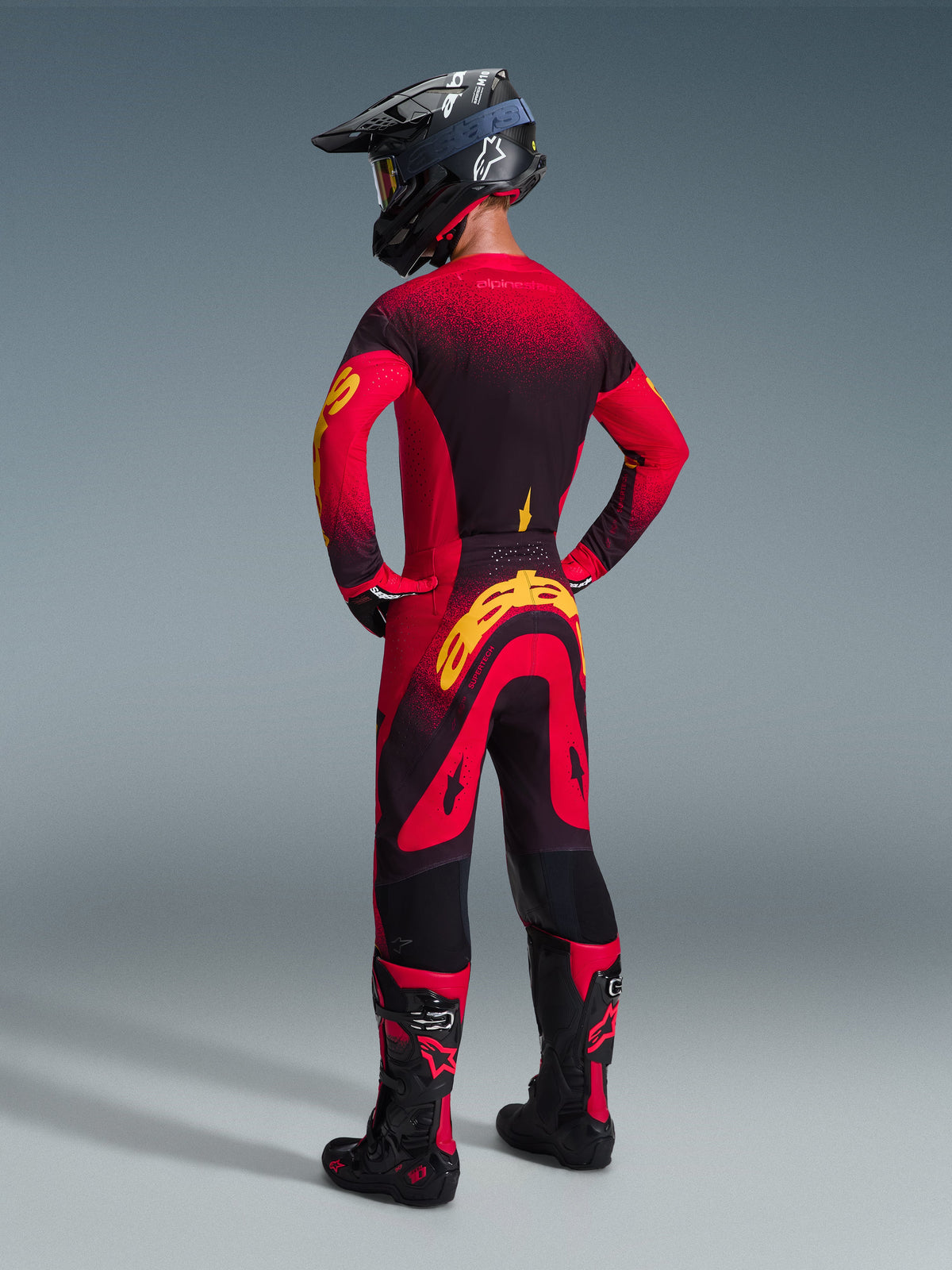 Piloto de motocross visto desde atrás vistiendo pantalones Alpinestars Supertech Scenz 2026 en rojo, negro y amarillo, con un gran logotipo amarillo en el asiento, combinado con un jersey a juego con degradado en rojo y negro, casco negro y botas de motocross rojas; equipamiento de competición profesional para rendimiento todoterreno.