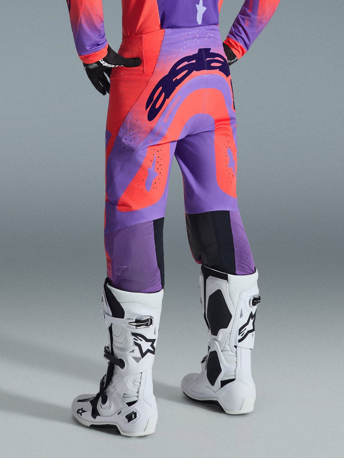 Pantalones Alpinestars Supertech Scenz 2026, pantalones de motocross, Hot Coral Purple, vista trasera con marca llamativa en el asiento, paneles vibrantes en coral y púrpura con zonas perforadas para ventilación, combinados con botas de carreras blancas y negras