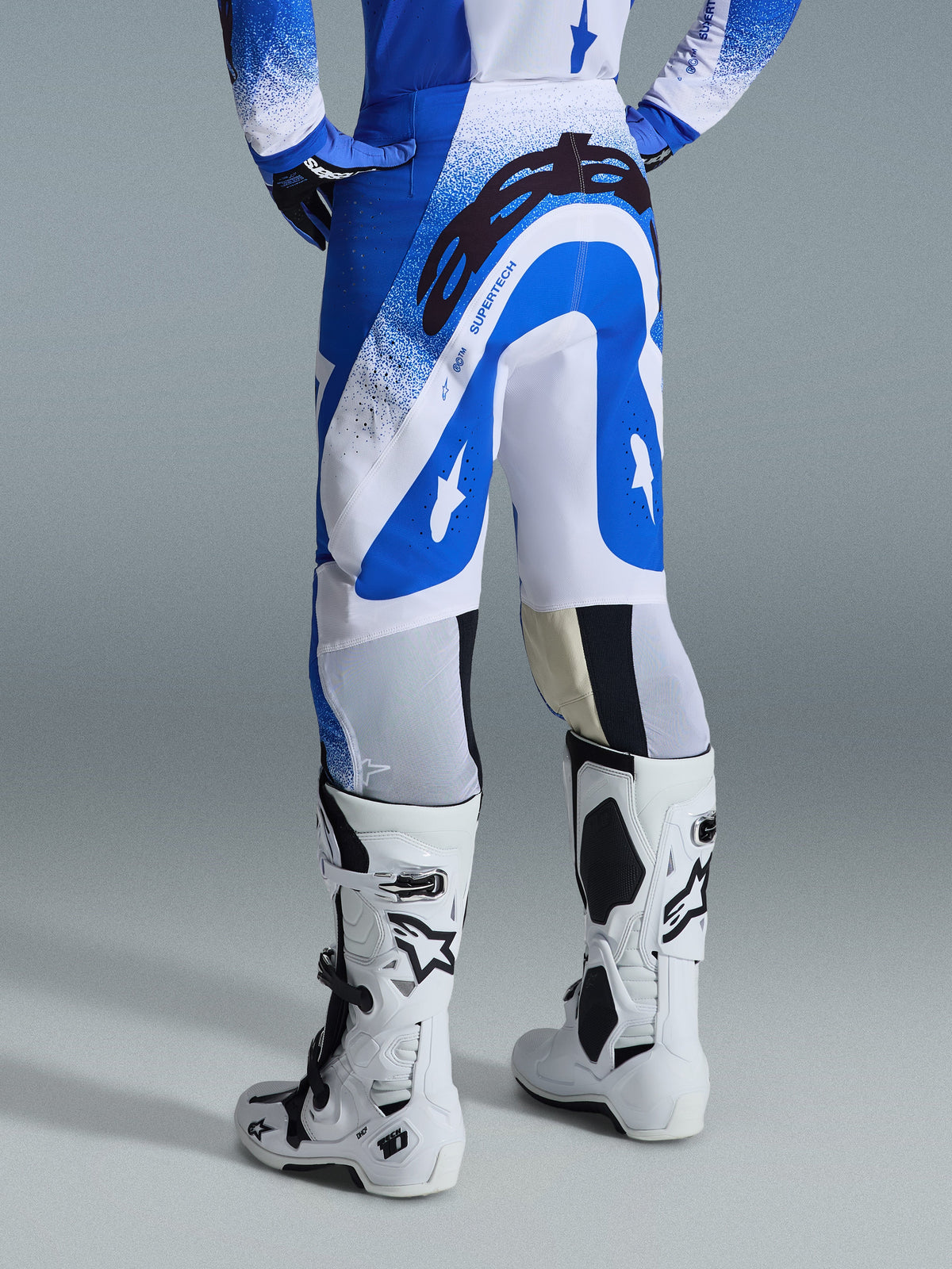Alpinestars 2026 Supertech Scenz Pants, pantalones de motocross, azul UCLA y blanco, vista trasera que muestra el gran logotipo negro de Astars en el asiento y la marca 'SUPERTECH', usados con botas Tech 10 blancas y un jersey azul y blanco a juego.