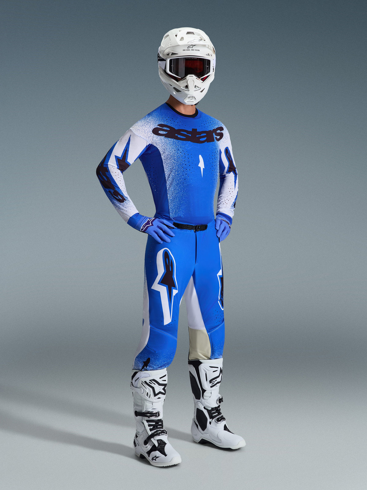 Piloto de motocross con equipamiento de carreras completo, que incluye los pantalones Supertech Scenz 2026 en azul Ucla y blanco, jersey a juego en azul y blanco con logotipo de Astars, casco blanco con gafas rojas y botas blancas y negras, conjunto coordinado para carreras profesionales de off-road.