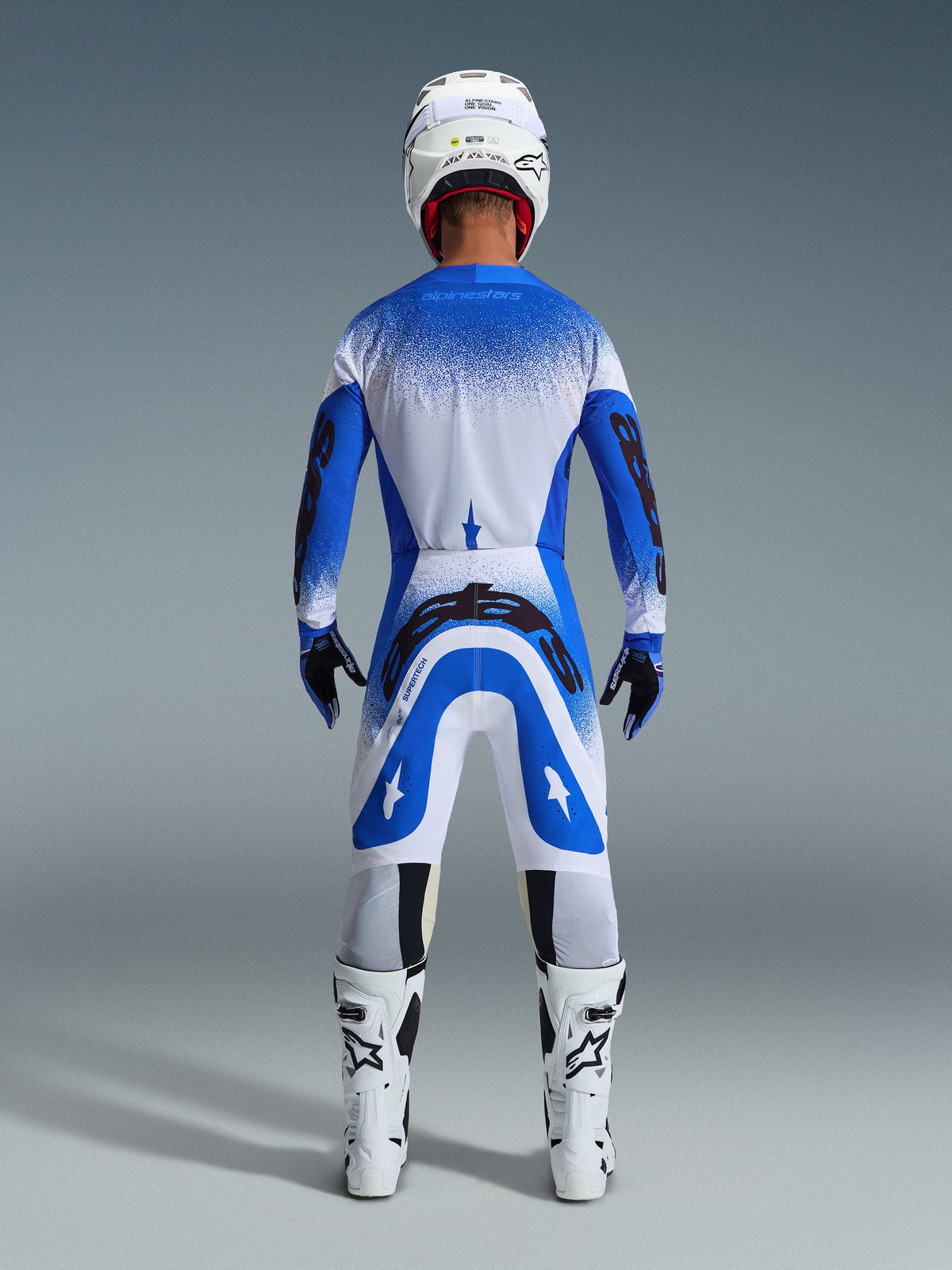 Piloto de motocross visto desde atrás con equipación completa de competición, pantalones Alpinestars 2026 Supertech Scenz en azul Ucla y blanco, jersey azul y blanco a juego con patrón moteado, casco blanco, botas blancas y negras, conjunto coordinado para carreras profesionales