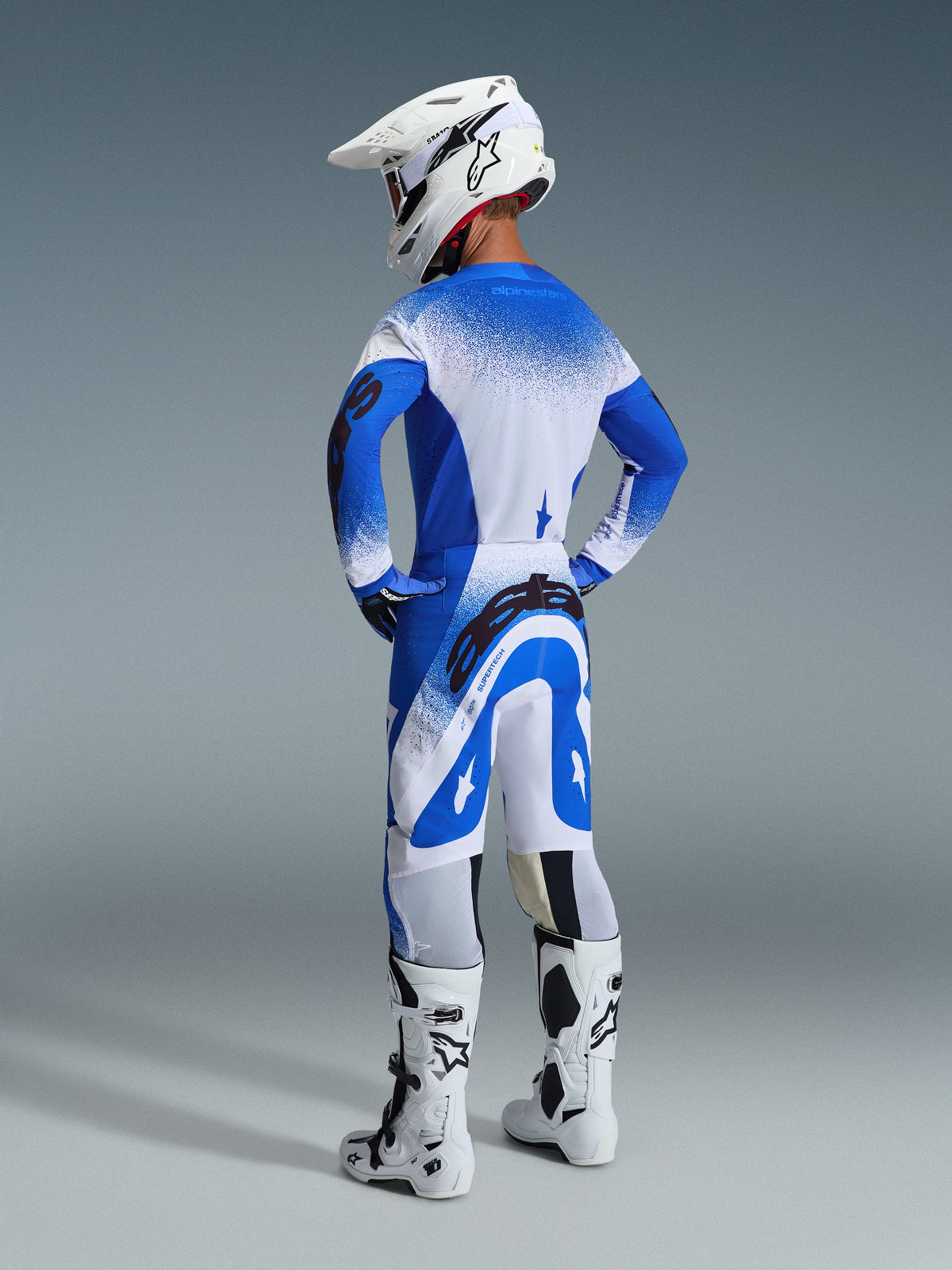 Piloto de motocross visto desde atrás con pantalones y jersey Supertech Scenz 2026 en azul Ucla y blanco, casco blanco con logo de Alpinestars, botas de motocross blancas y negras, equipo profesional de carreras off-road.