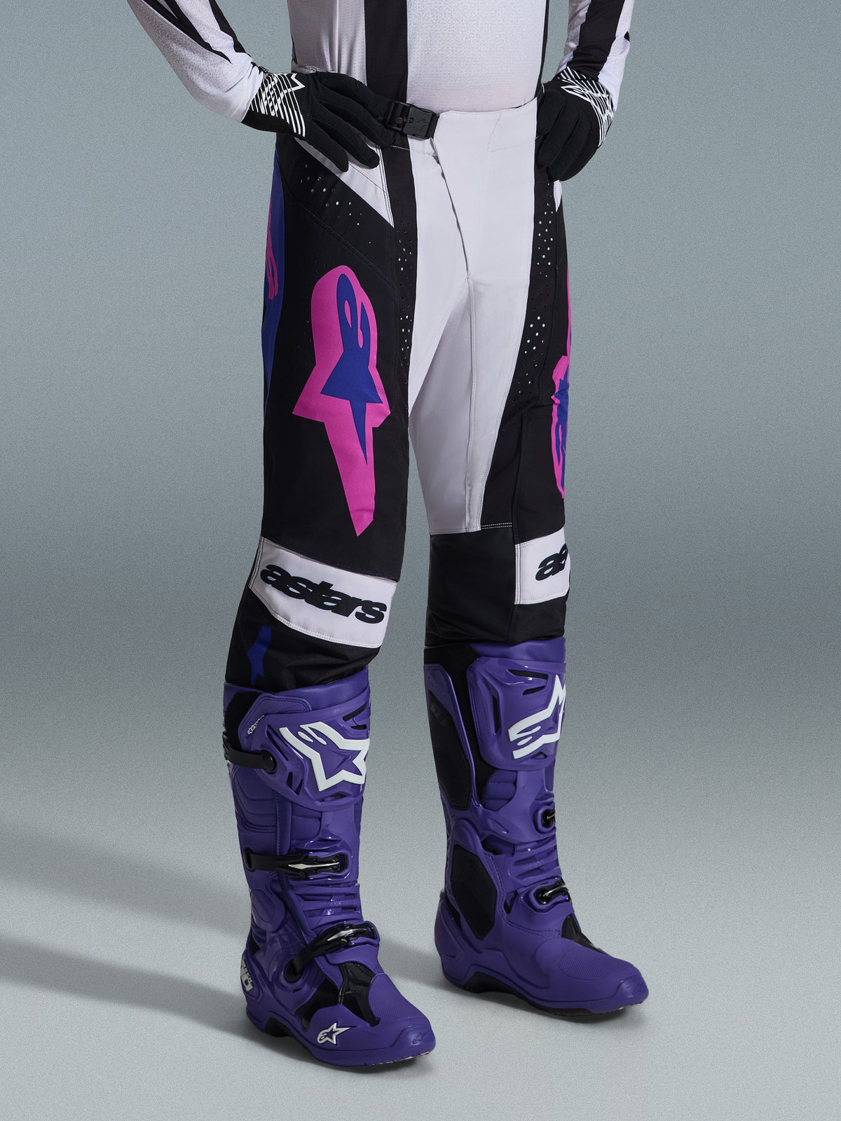 Pantalones Alpinestars Techstar Knif 2026, en color negro, gris claro y morado, combinados con botas de motocross moradas a juego, con llamativos detalles en rosa y marca 'astars', equipamiento profesional para carreras de motocross