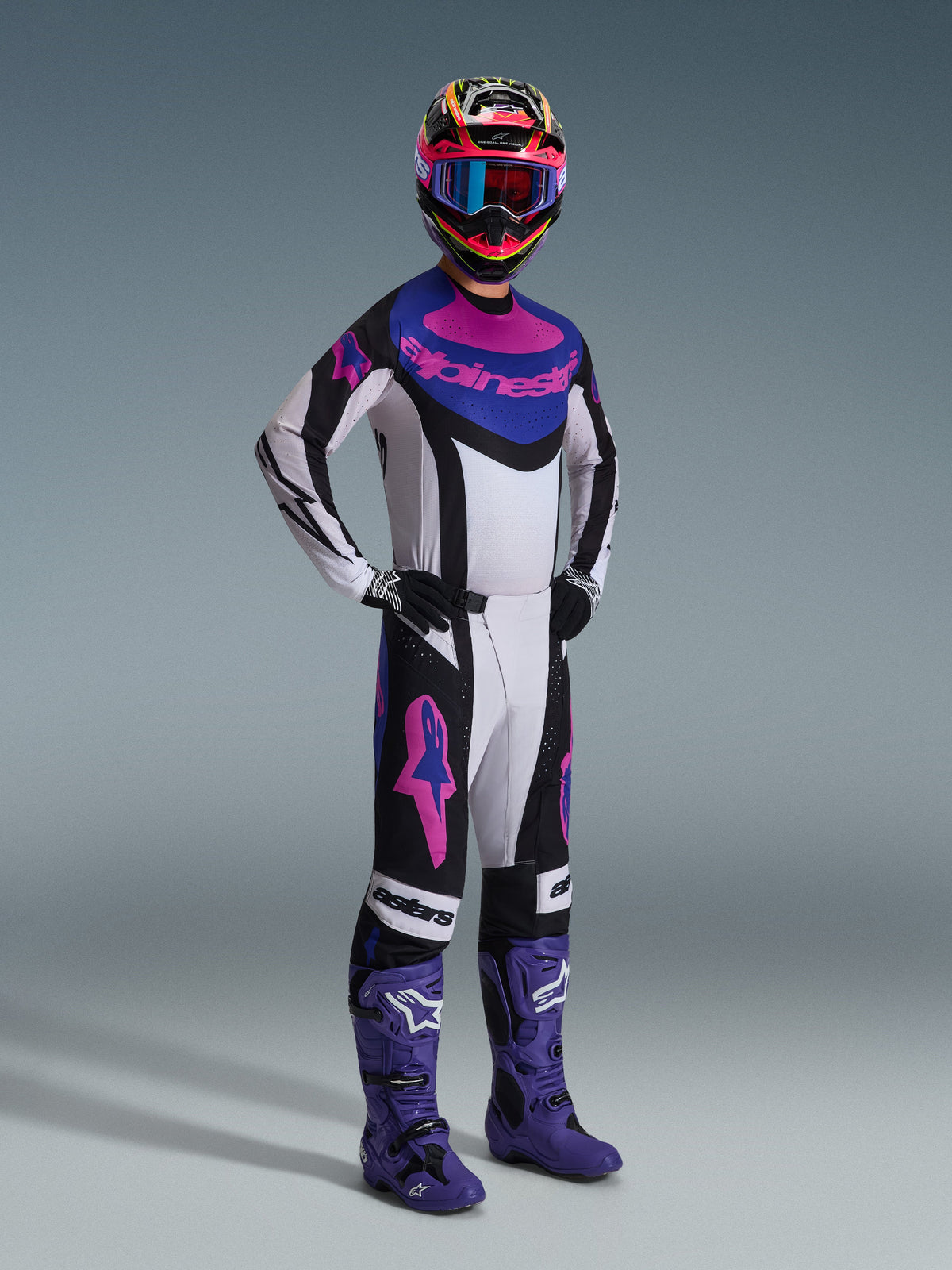 Motocross rider wearing the Alpinestars 2026 Techstar Knif Gear Set, white jersey with black, blue, and pink accents, matching pants with pink Astars logos, multi-colored helmet with goggles, purple racing boots, coordinated high-performance gear for motocross competition
