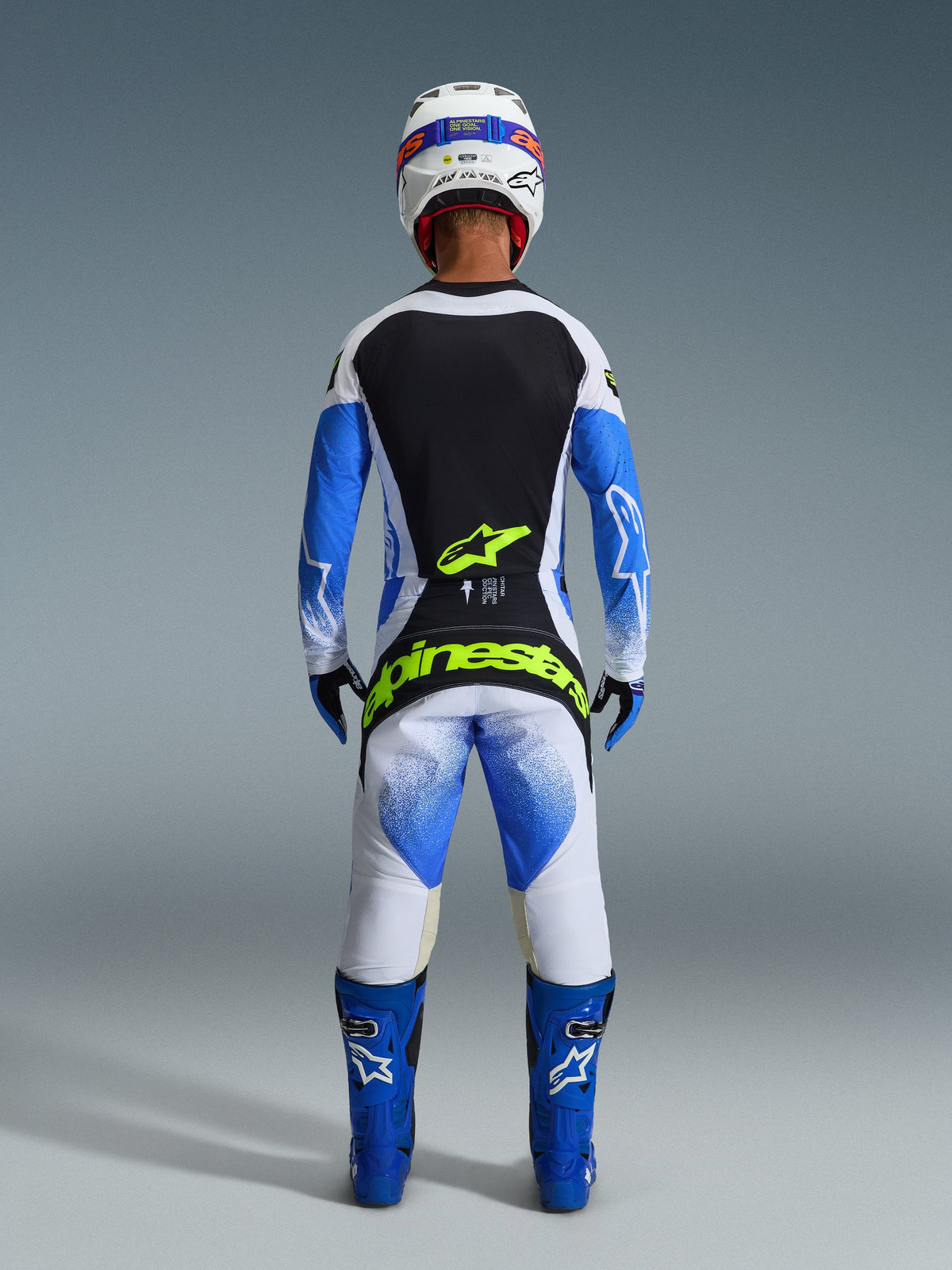 Piloto de motocross de espaldas con equipamiento de carreras completo, pantalones Techstar Knif 2026 en UCLA azul, negro y amarillo flúor, jersey de manga larga a juego con paneles negros y blancos, botas azules, casco blanco y el logotipo de Alpinestars en amarillo flúor en el asiento.