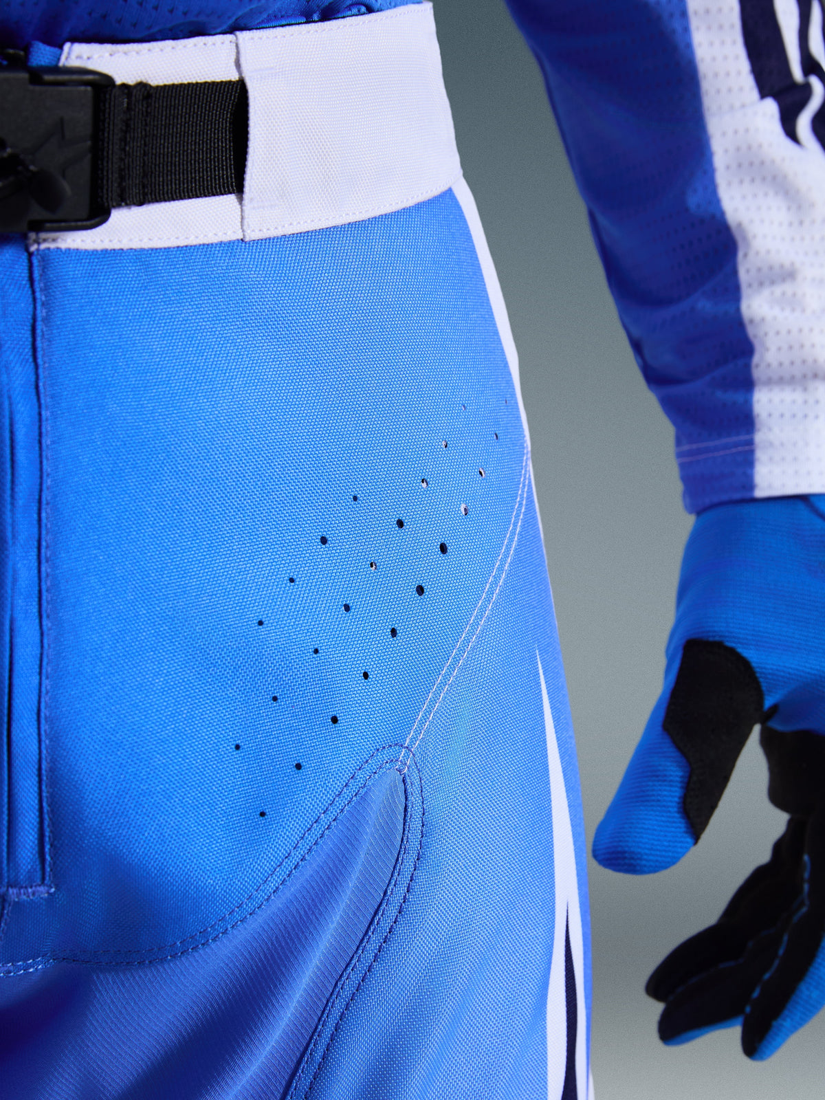 Alpinestars Techstar Knif Pants 2026, azul UCLA y blanco, primer plano de la zona de la cadera con perforaciones láser para ventilación, cierre de cintura con hebilla magnética SX2 y guantes de motocross a juego