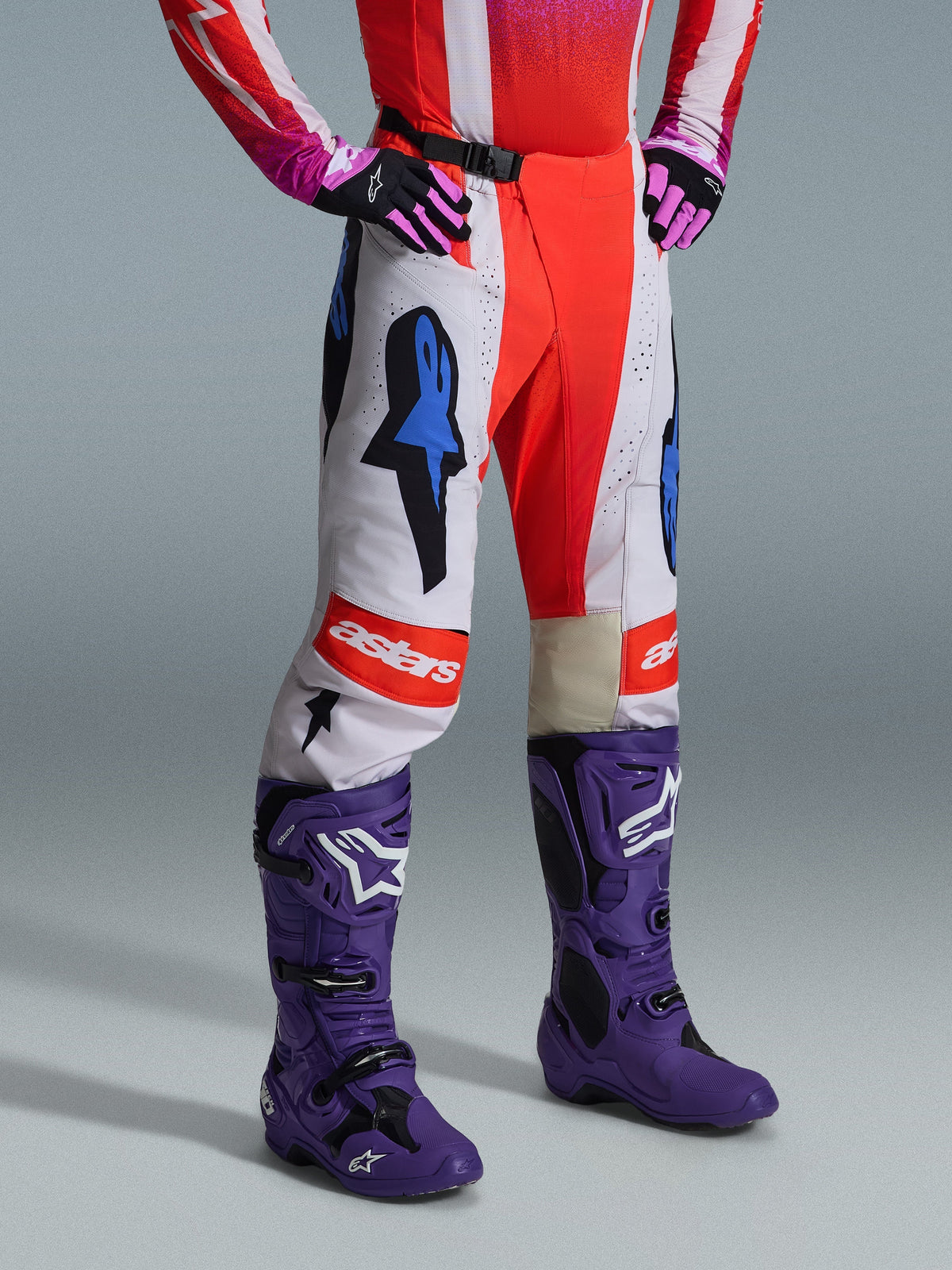 Alpinestars 2026 Techstar Knif Pants, pantalones de motocross, blancos y rojo brillante con acentos gráficos azules, con el logo "astars" en la rodilla, combinados con botas moradas y guantes negros y rosas, conjunto de equipamiento profesional para carreras off-road.