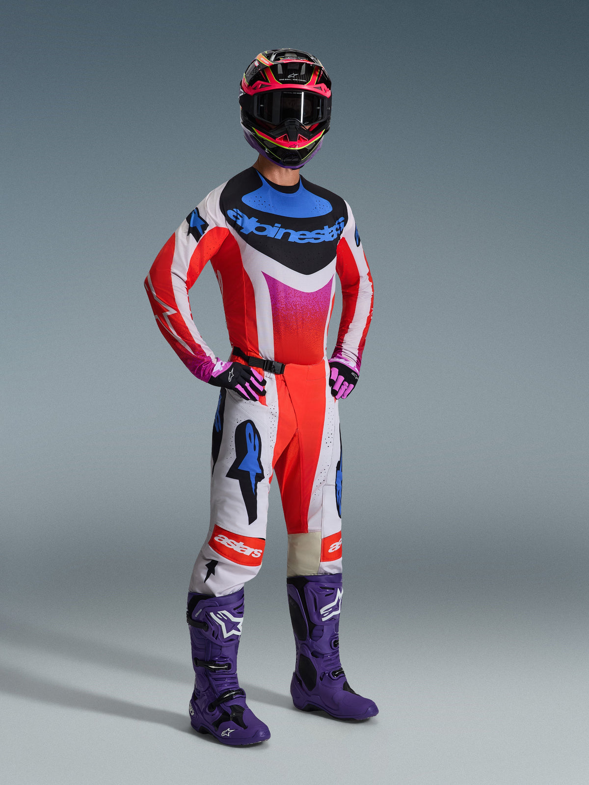 Piloto de motocross con equipamiento completo de Alpinestars, Jersey Techstar Knif 2026 en blanco y rojo con detalles azules y rosas, pantalones de motocross a juego, botas moradas y casco multicolor, conjunto profesional coordinado para rendimiento todoterreno