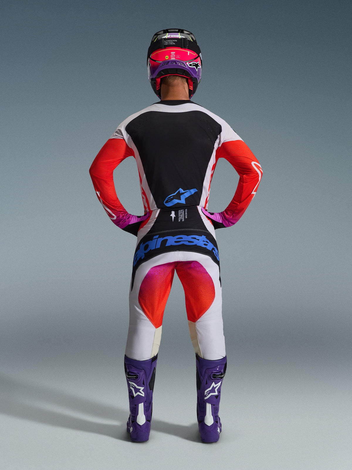 Piloto de motocross visto desde atrás con pantalones Techstar Knif 2026 y jersey a juego, diseño gris claro y negro con detalles en naranja y púrpura, logotipo azul de Alpinestars en el asiento, casco y botas púrpuras, conjunto de equipamiento profesional para carreras off-road.