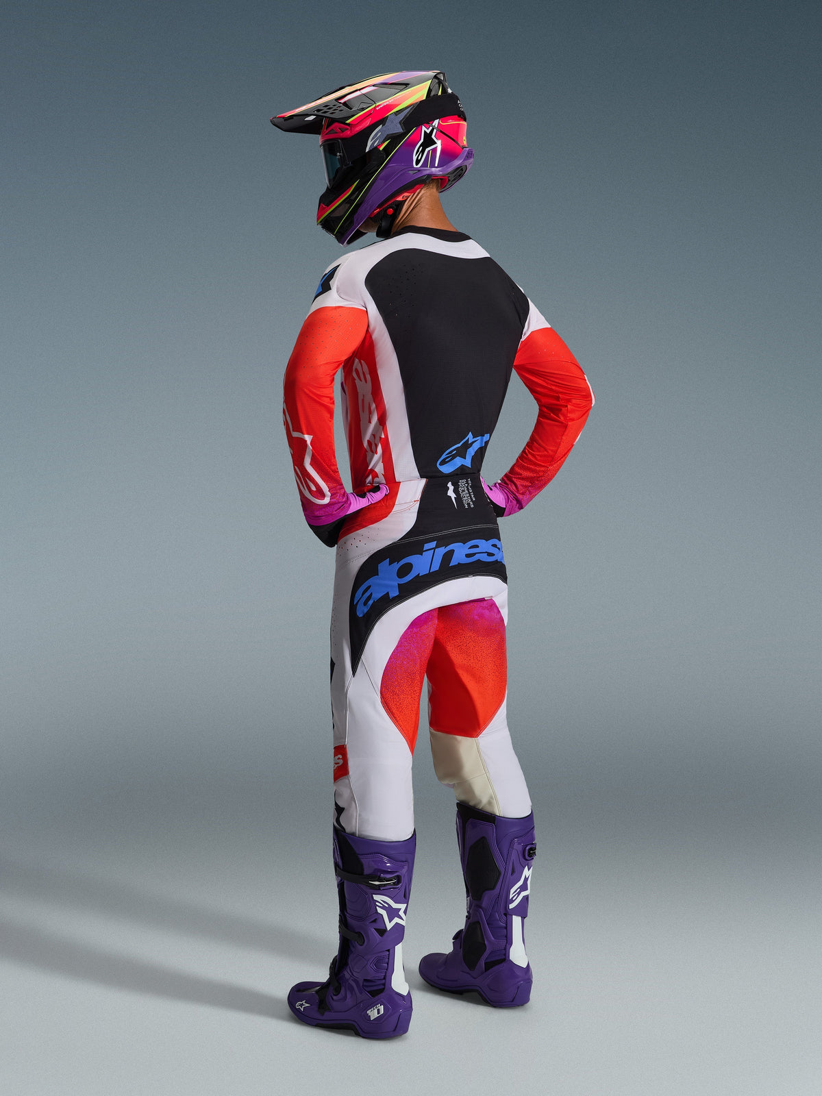 Piloto de motocross visto desde atrás con equipamiento completo, pantalones Alpinestars Techstar Knif 2026 en gris claro y naranja flúor con logotipos azules, jersey a juego con mangas naranjas, botas Tech 10 moradas y casco de carreras colorido, conjunto profesional coordinado para off-road