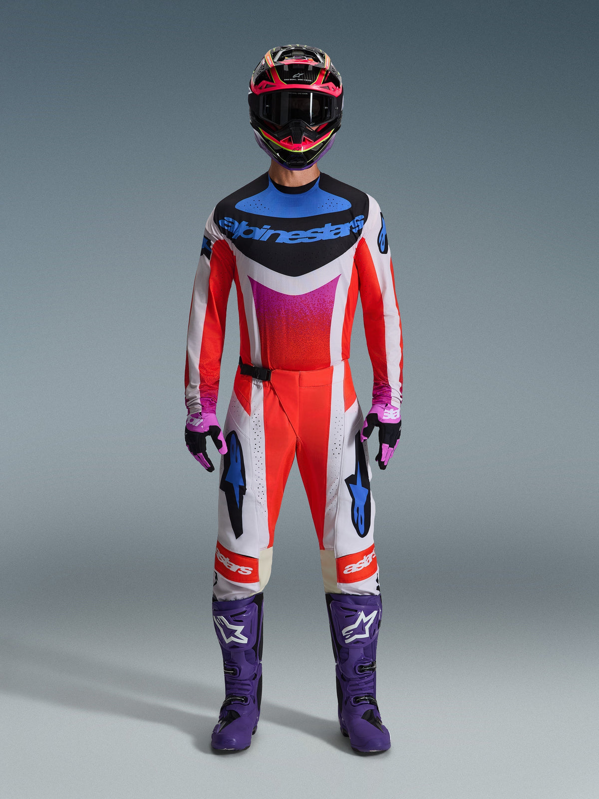 Piloto de motocross con equipamiento completo de competición, pantalones Alpinestars 2026 Techstar Knif, diseño naranja flúor y blanco con acentos azules, jersey multicolor a juego, botas moradas, guantes negros y morados, traje profesional coordinado para motocross