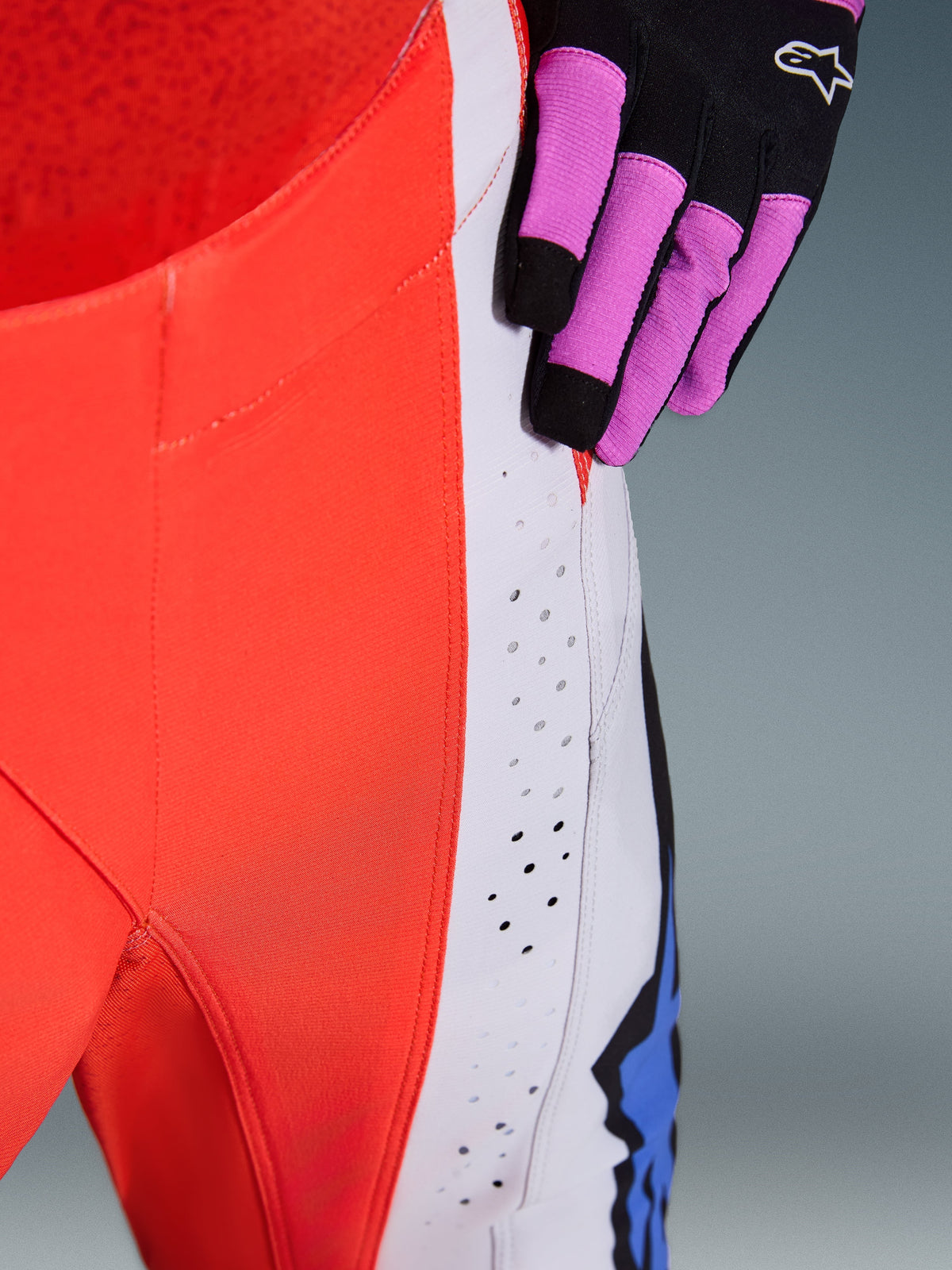 Pantalones Alpinestars Techstar Knif 2026, pantalones de motocross, naranja flúor y gris claro con detalles azules, primer plano que muestra las perforaciones de ventilación cortadas con láser en el muslo, usados con un guante de motocross negro y púrpura