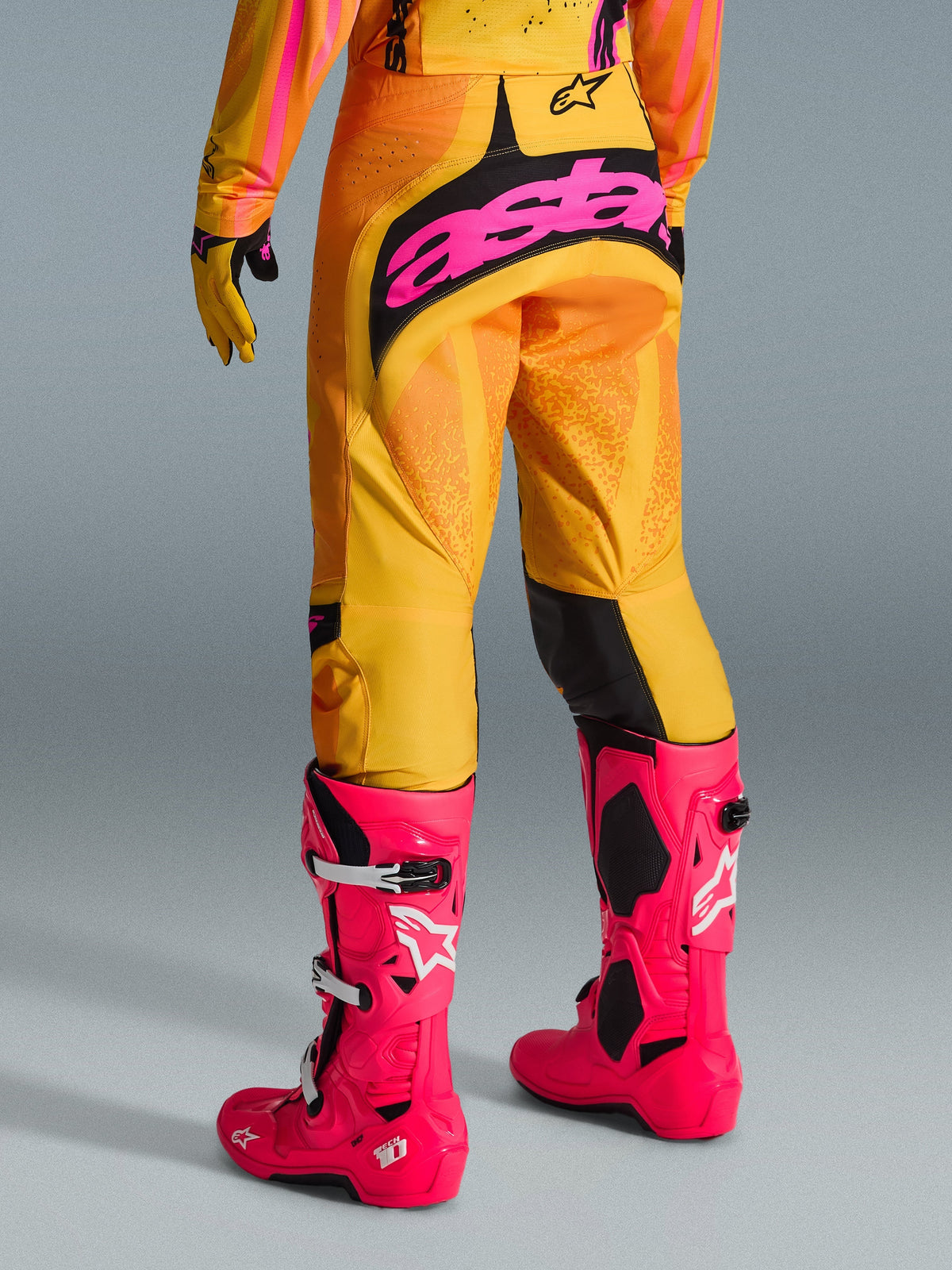 Pantalones Alpinestars Techstar Nomur 2026, pantalones de motocross, naranja, amarillo y rosa, vista trasera con el gran logotipo 'astars' en rosa sobre el asiento, combinados con botas Tech 10 rosa brillante y jersey a juego, equipamiento de alto rendimiento para motocross