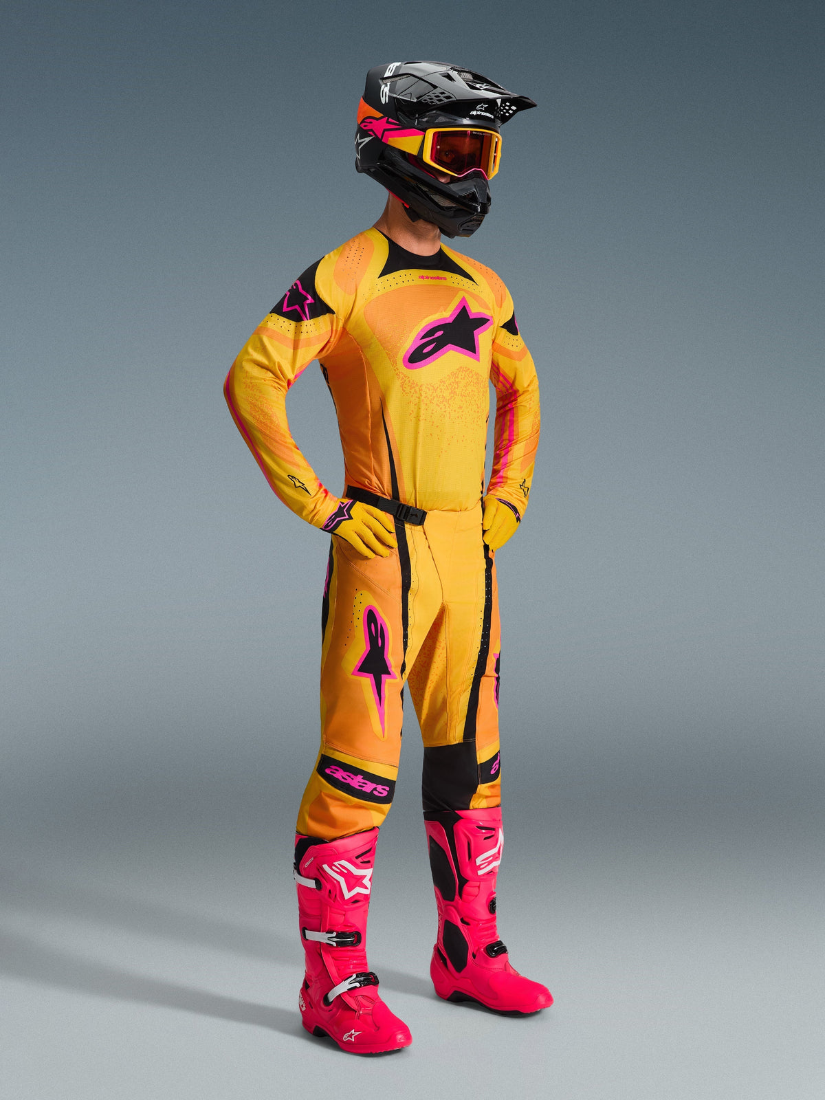 Piloto de motocross con pantalones Alpinestars Techstar Nomur 2026 en naranja, amarillo y rosa, equipo de carreras completo con jersey y guantes a juego, botas rosa brillante, casco negro con gafas naranjas, vista de cuerpo completo sobre fondo gris, traje profesional de competición off-road