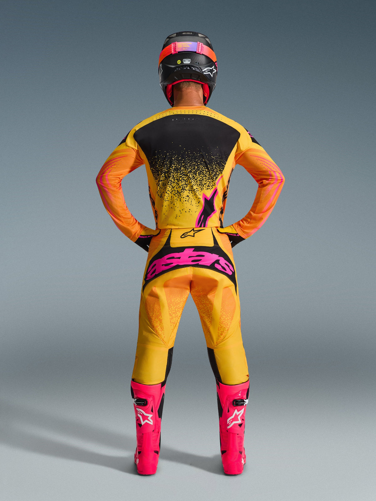 Vista posterior de un piloto de motocross con los pantalones Alpinestars 2026 Techstar Nomur en naranja, amarillo y rosa, con un gran logotipo rosa en el asiento, combinados con una camiseta amarilla y negra a juego, botas rosa brillante y un casco negro y rosa.