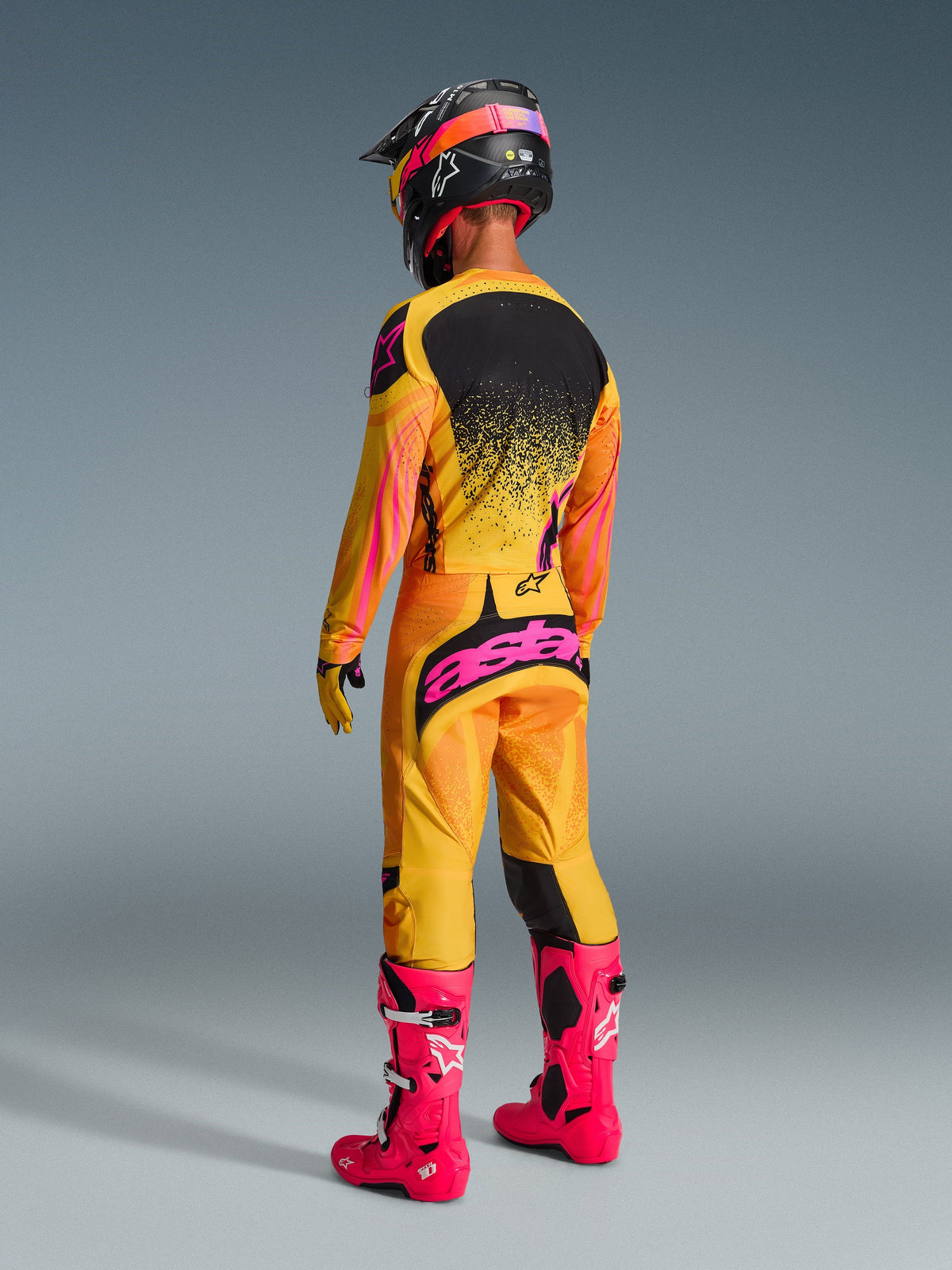 Piloto de motocross visto desde atrás con equipo de competición completo, pantalones Techstar Nomur 2026 en naranja, amarillo y rosa, logotipo 'astars' rosa llamativo en el asiento, jersey a juego, botas rosa brillante y casco negro con detalles en rosa, conjunto coordinado para competición todoterreno.