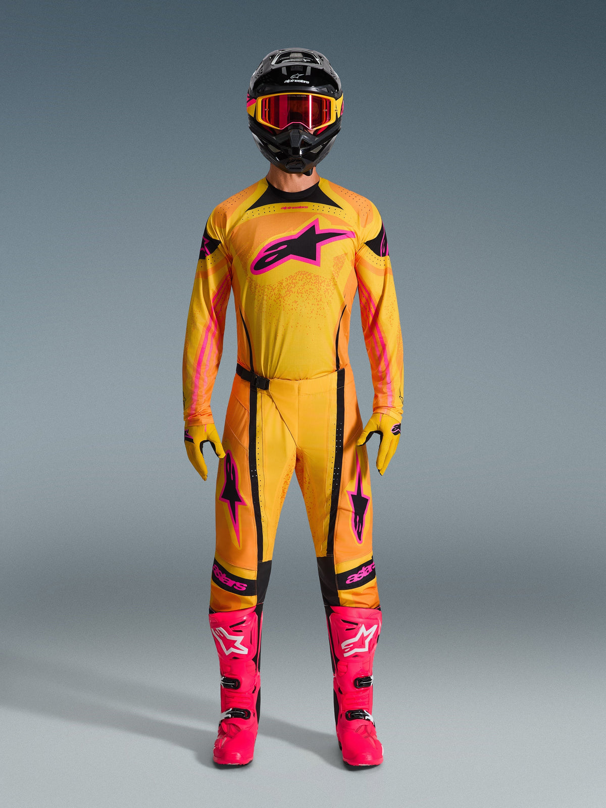 Piloto de motocross con conjunto completo Alpinestars 2026 Techstar Nomur, jersey y pantalones en naranja, amarillo y rosa, guantes amarillos y rosas a juego, botas de competición en rosa vibrante y casco negro con gafas rosas, atuendo de motocross de alto rendimiento coordinado.